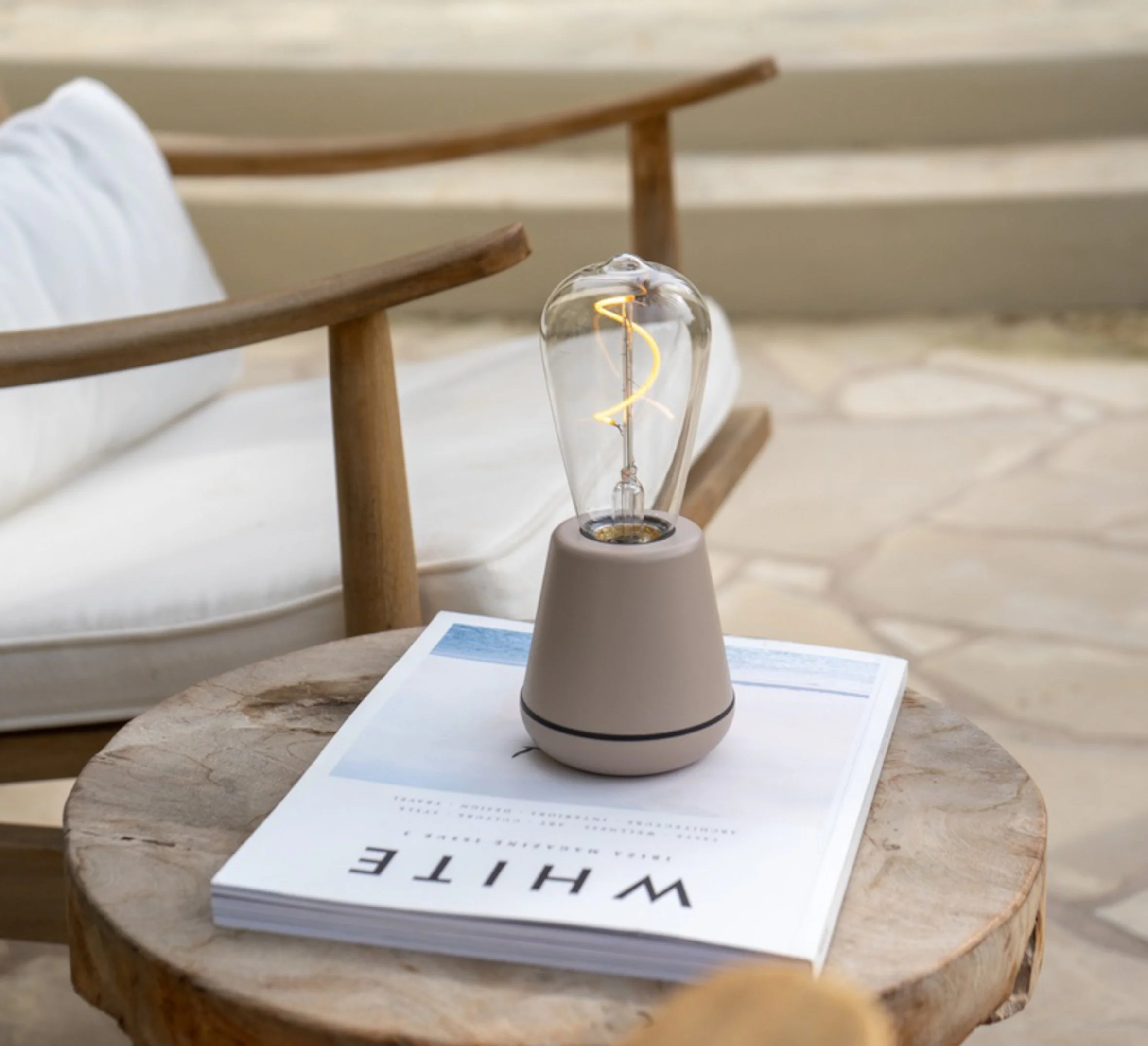 an outdoor lamp with a beige base and bulb on top with no lampshade sitting on a magazine on a wooden table next to a wooden chair with white upholstery
