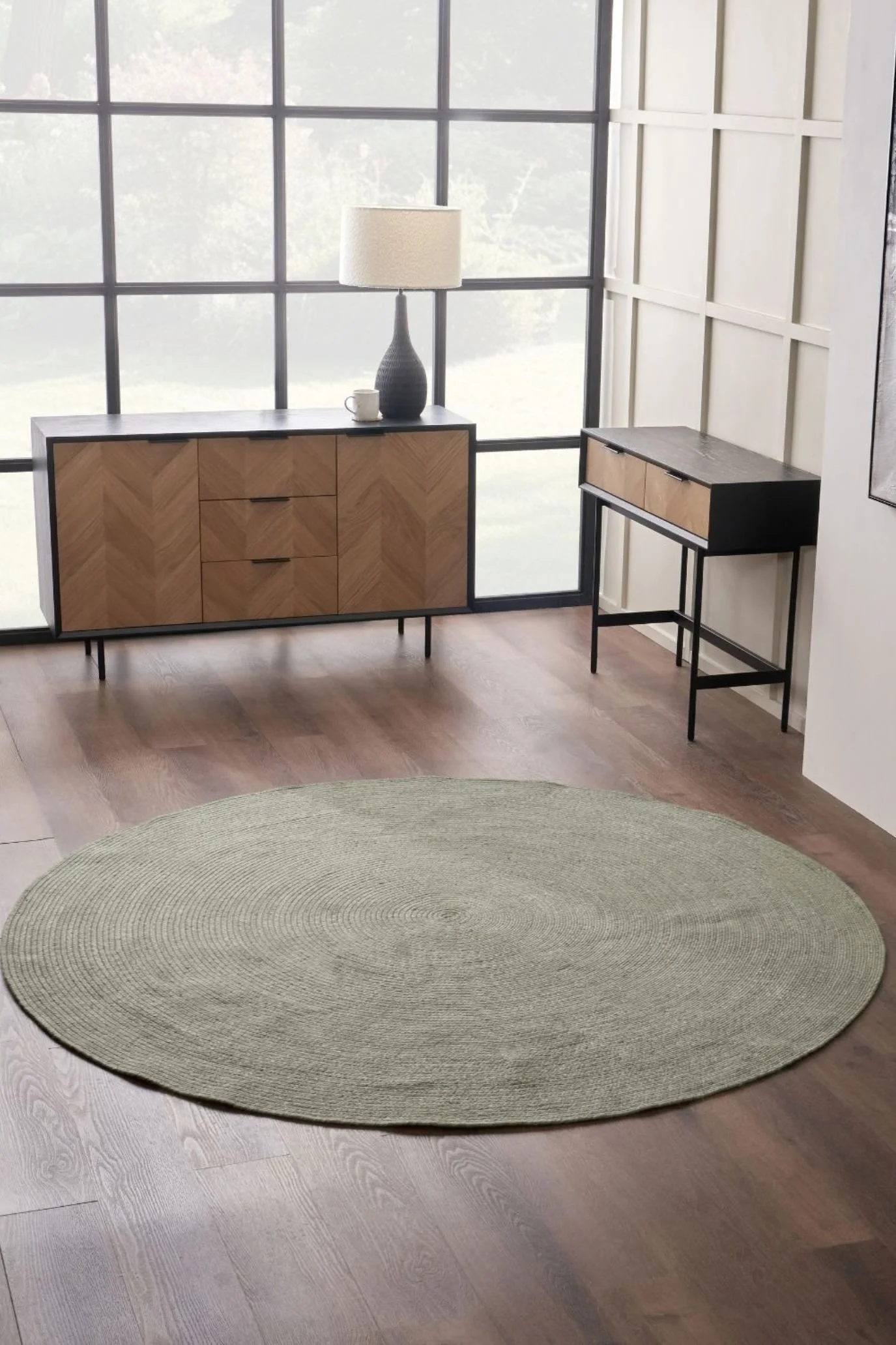 Round sage green rug on a wooden floor, with a sideboard and desk beside large windows.