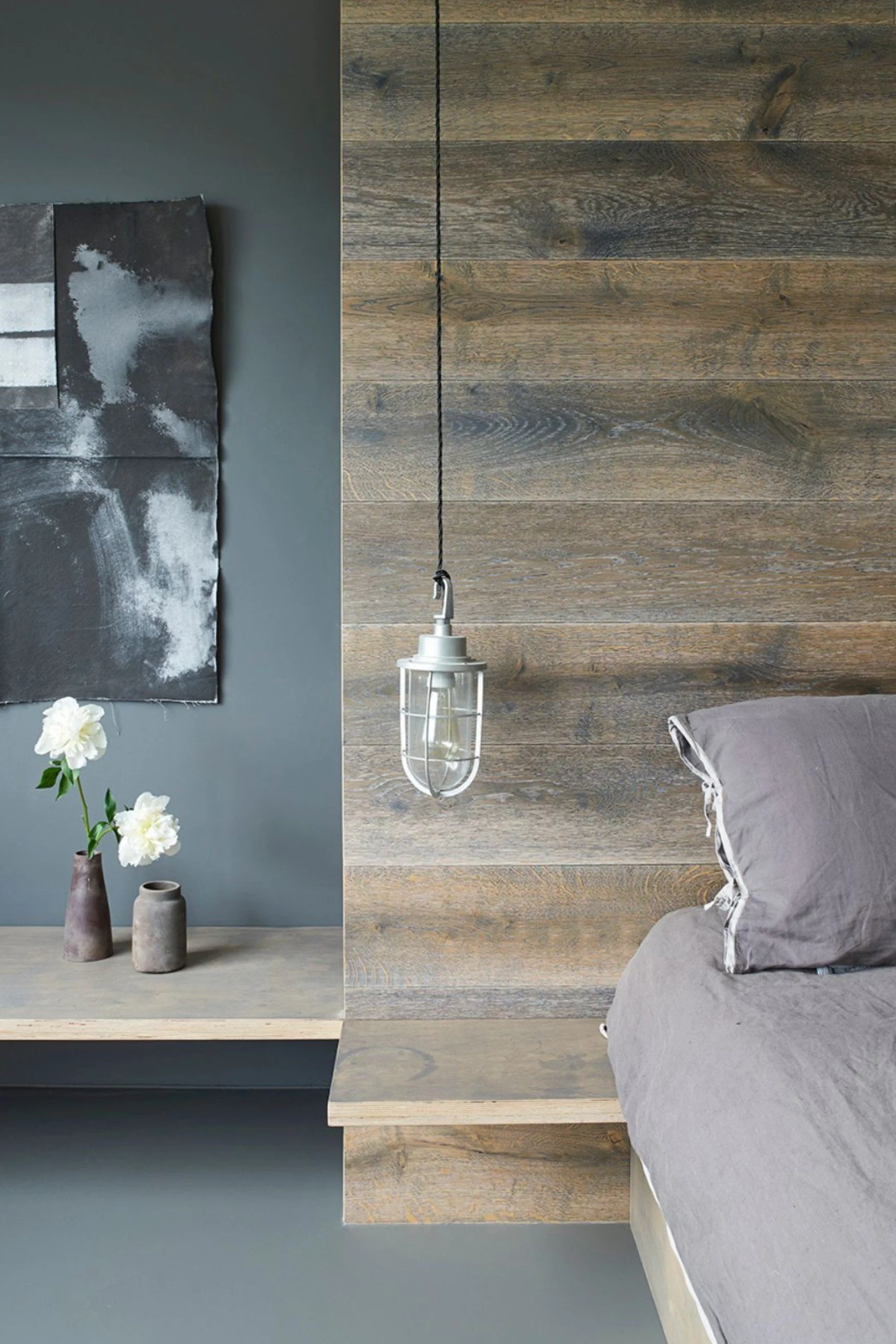A bed in grey linen infront of a custom made timber headboard with a hanging lamp beside it. There is black and white artwork on the wall and a shelf with white flowers in grey post