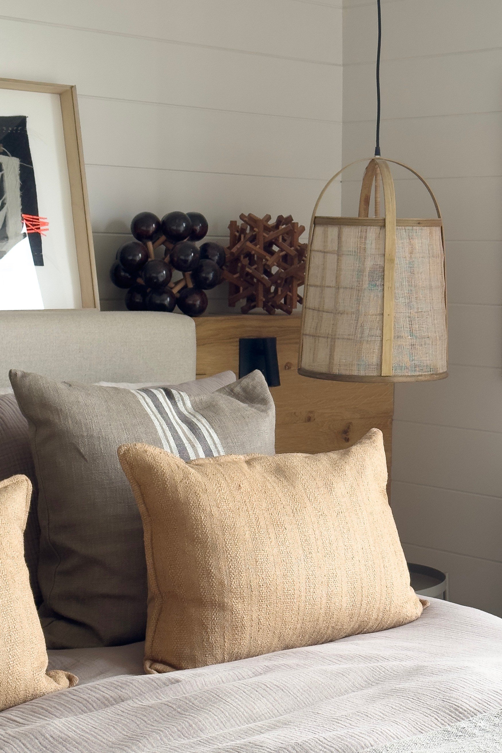A bed with a grey headboard against wooden shelving with a painting and two sculptural objects on display with beige and a yellow cushion and rattan lights hanging either side