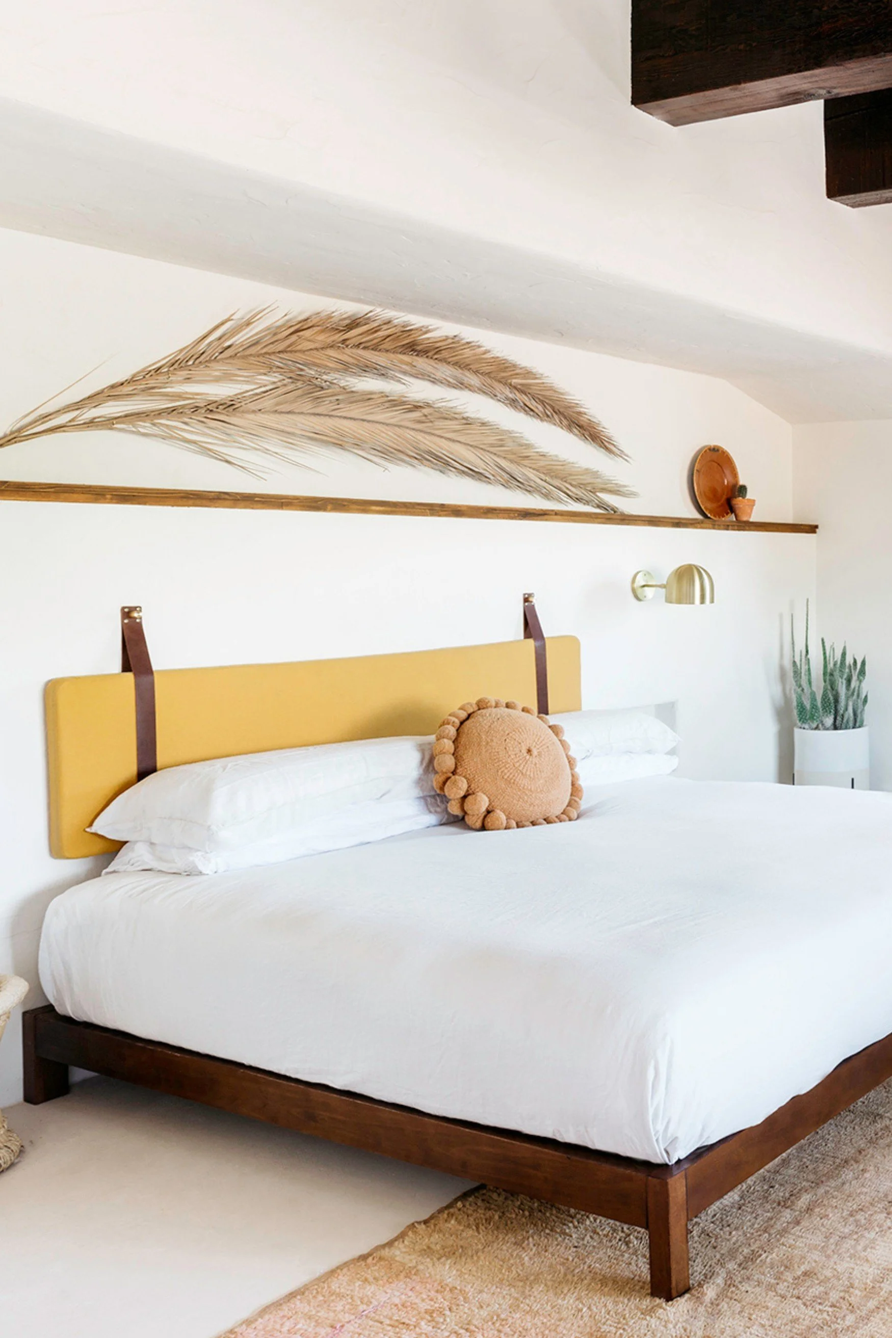 A bedroom with a yellow fabric headboard suspended by leather straps above a bed dressed in white with a huge dried palm leaf on a shelf above it