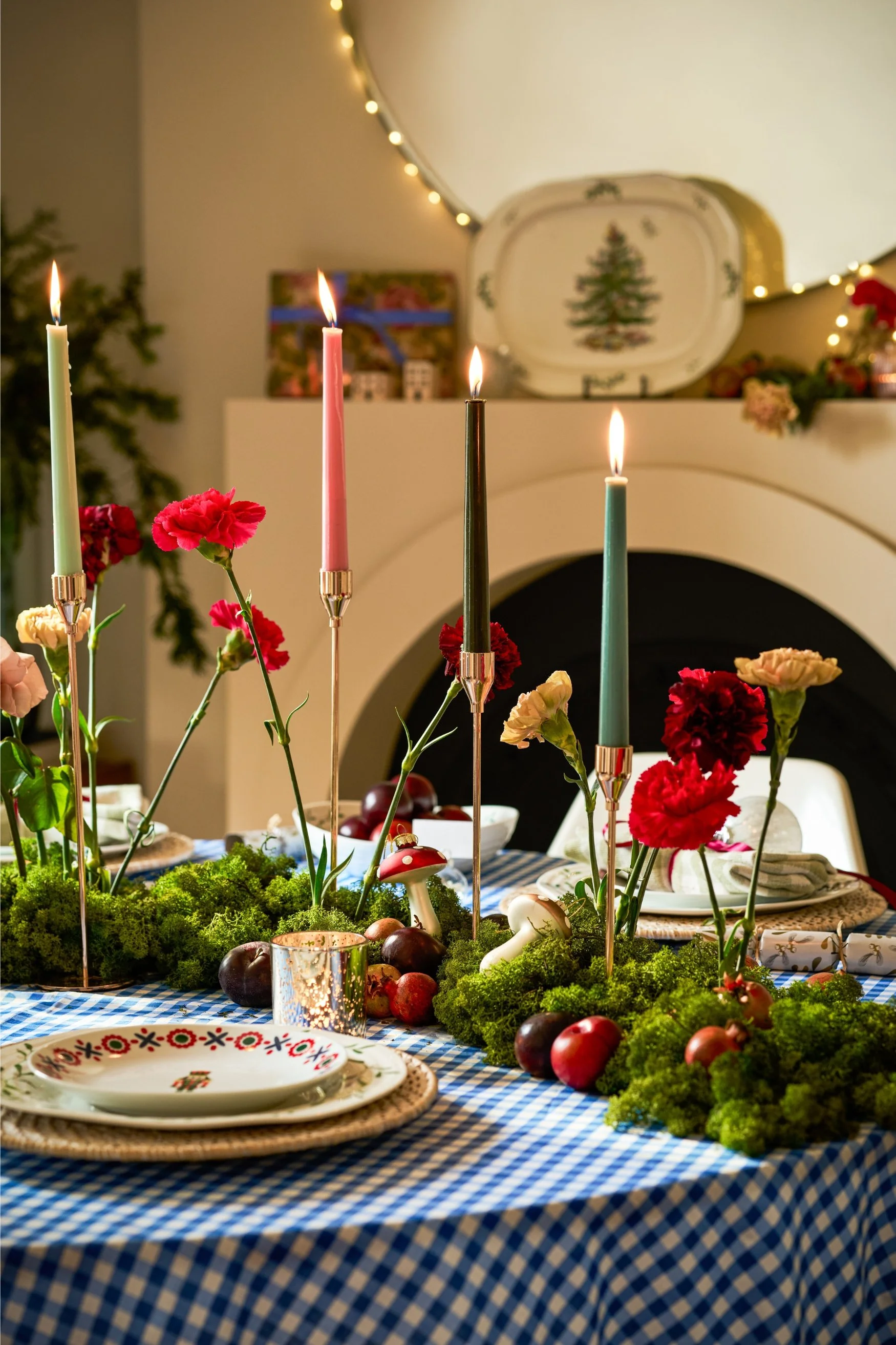 A table with a checkered blue cloth and four candle holders with lit candles and flowers poked into greenery