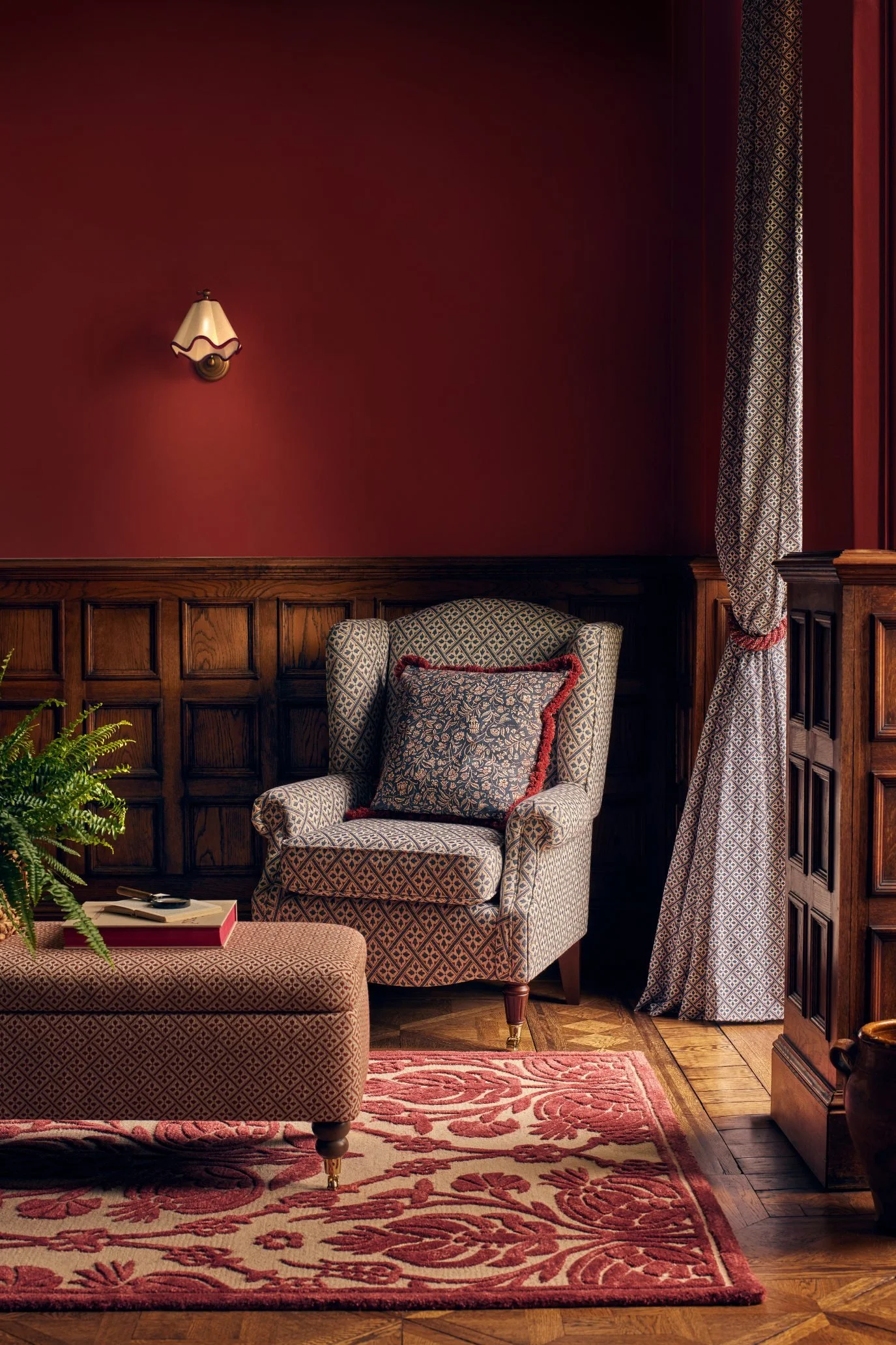 Living room with deep red accessories, floral patterned rug, fringed cushion and warm dark wood panelling.
