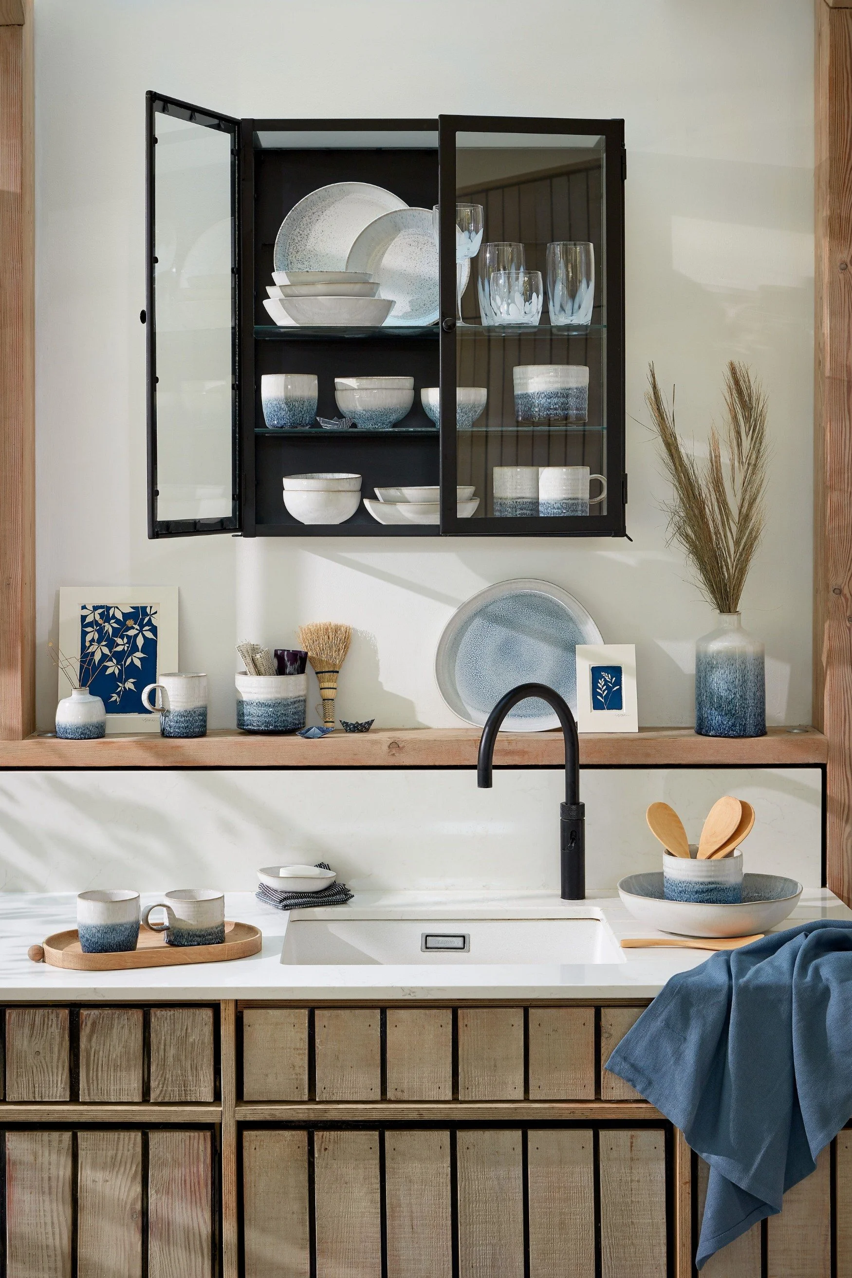 A sink with a black tap and white worktop  and black glass and metal cabinet all with blue and white glazed cups, bowls, plates and other vessels