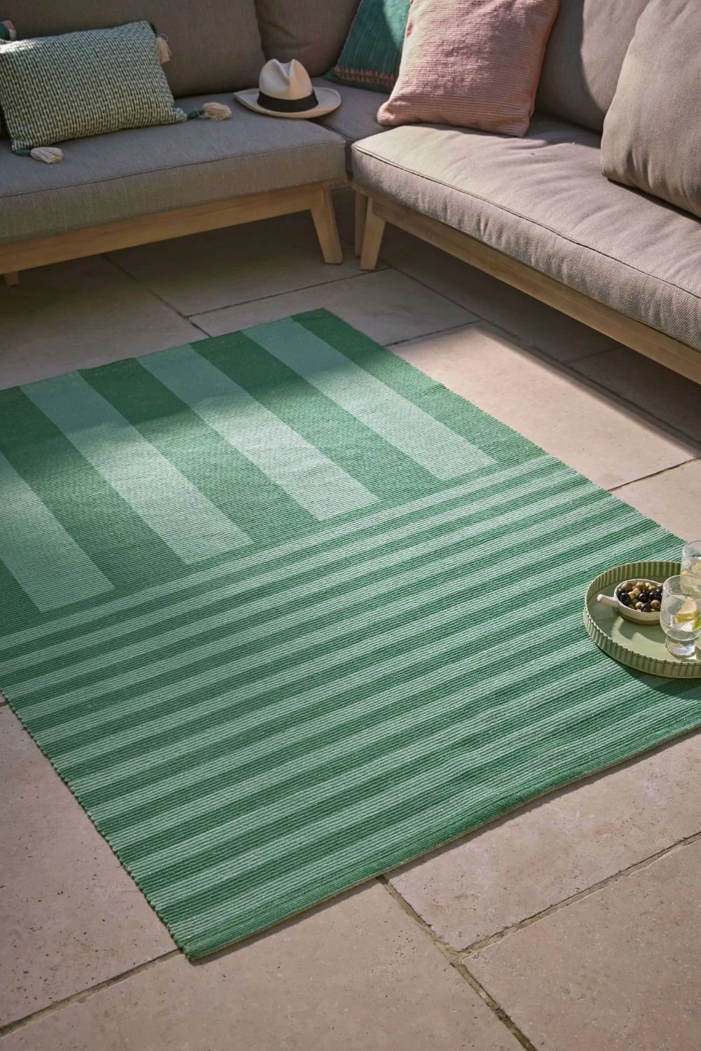 a green tonal rug it both wide and narrow stripes next to a wood and linen outdoor sofa