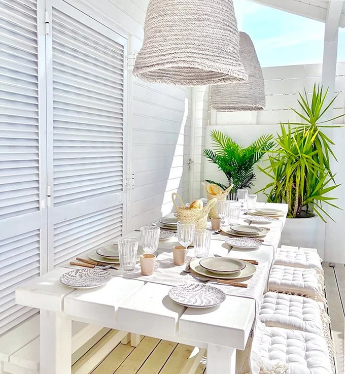 An outdoor white table with tableware, linen runners and glasses with two large rope pendant lights hanging above and potted plants behind
