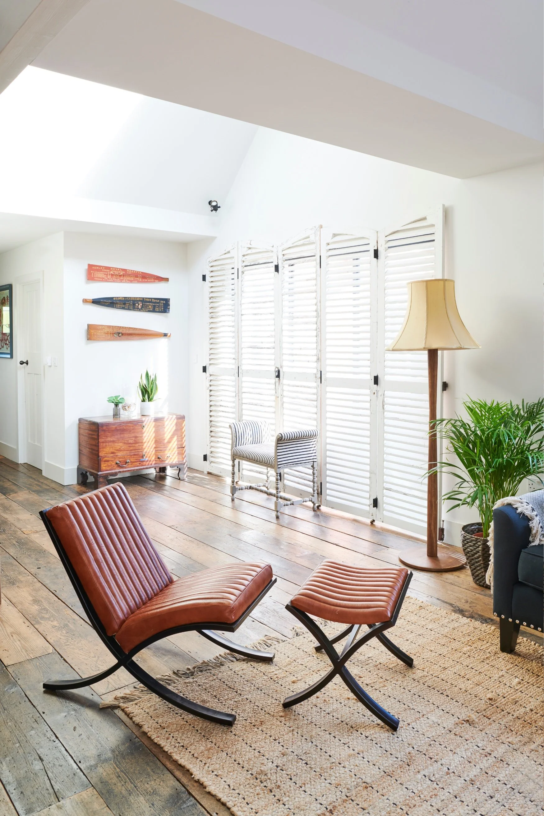 A retro style leather chair on a salvaged wooden floor in an open plan area with vintage shutters, a vintage brown wood chest with three old oars above it