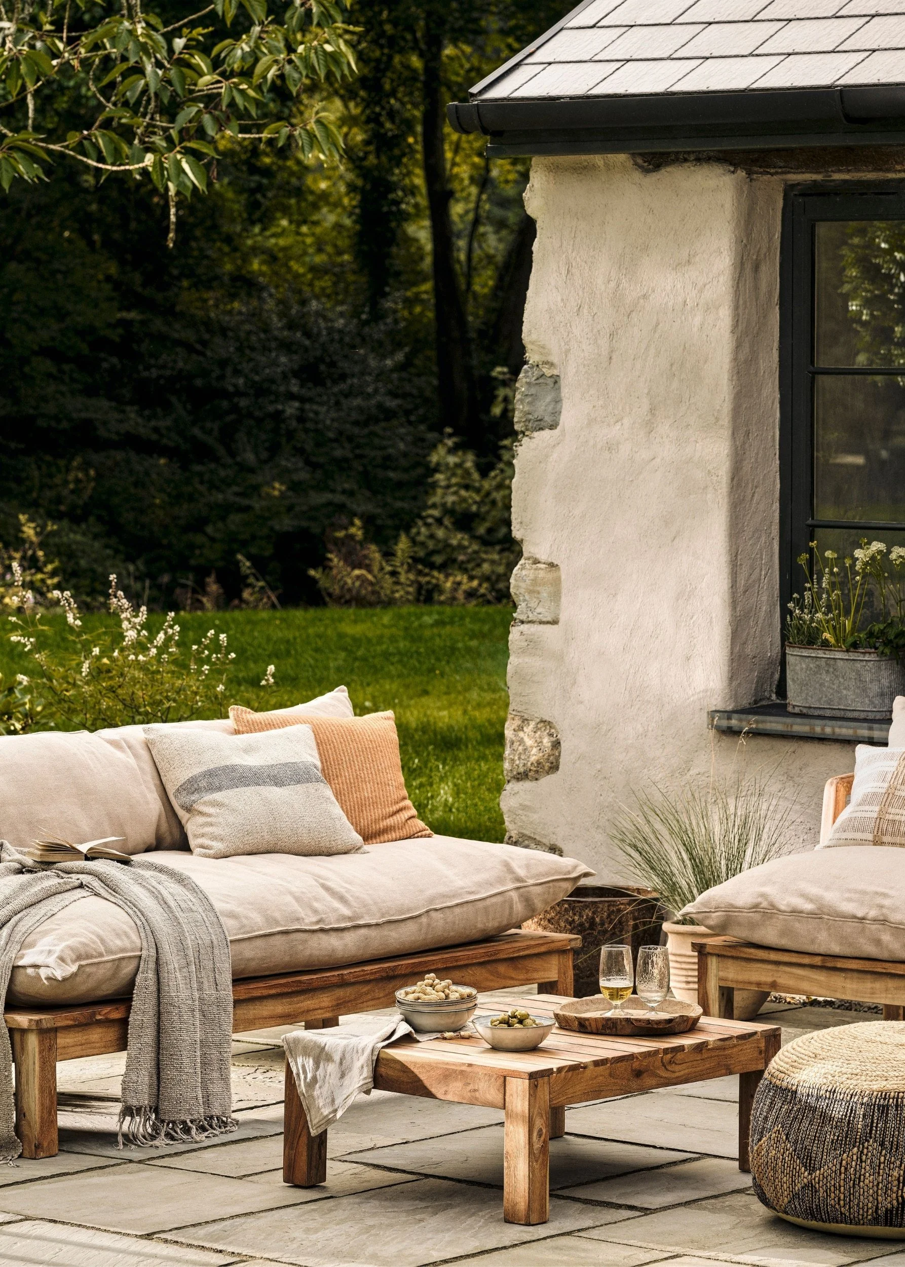 An outdoor sofa and coffee table made of wood with cream seating and cushions in different colours infront of a garden and traditional looking building on a tiled patio