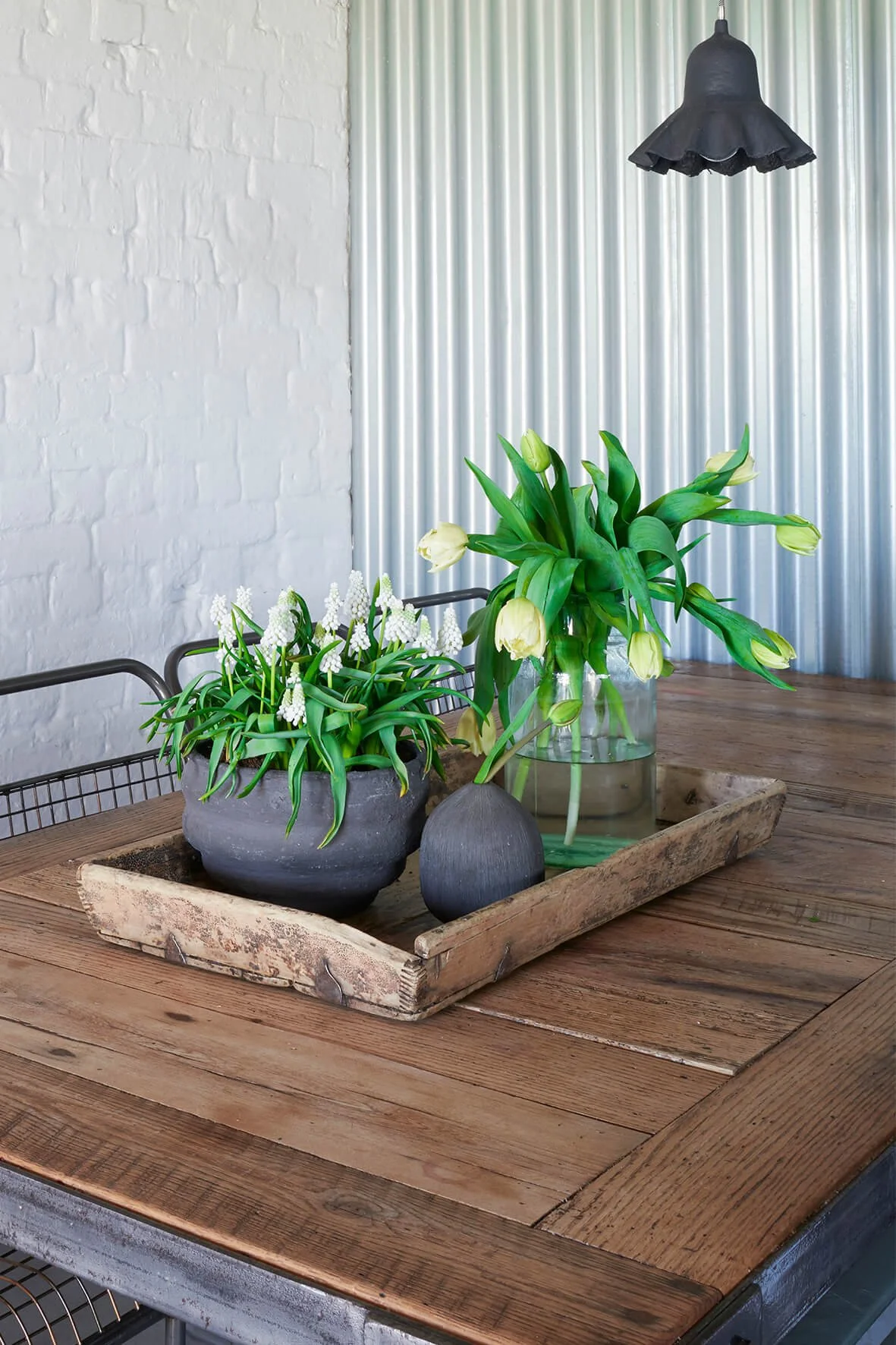 a wooden tray on a wooden table with a grey pot of white grape hyacinths and a vase of tulips with a corrugated iron wall and black pendant light