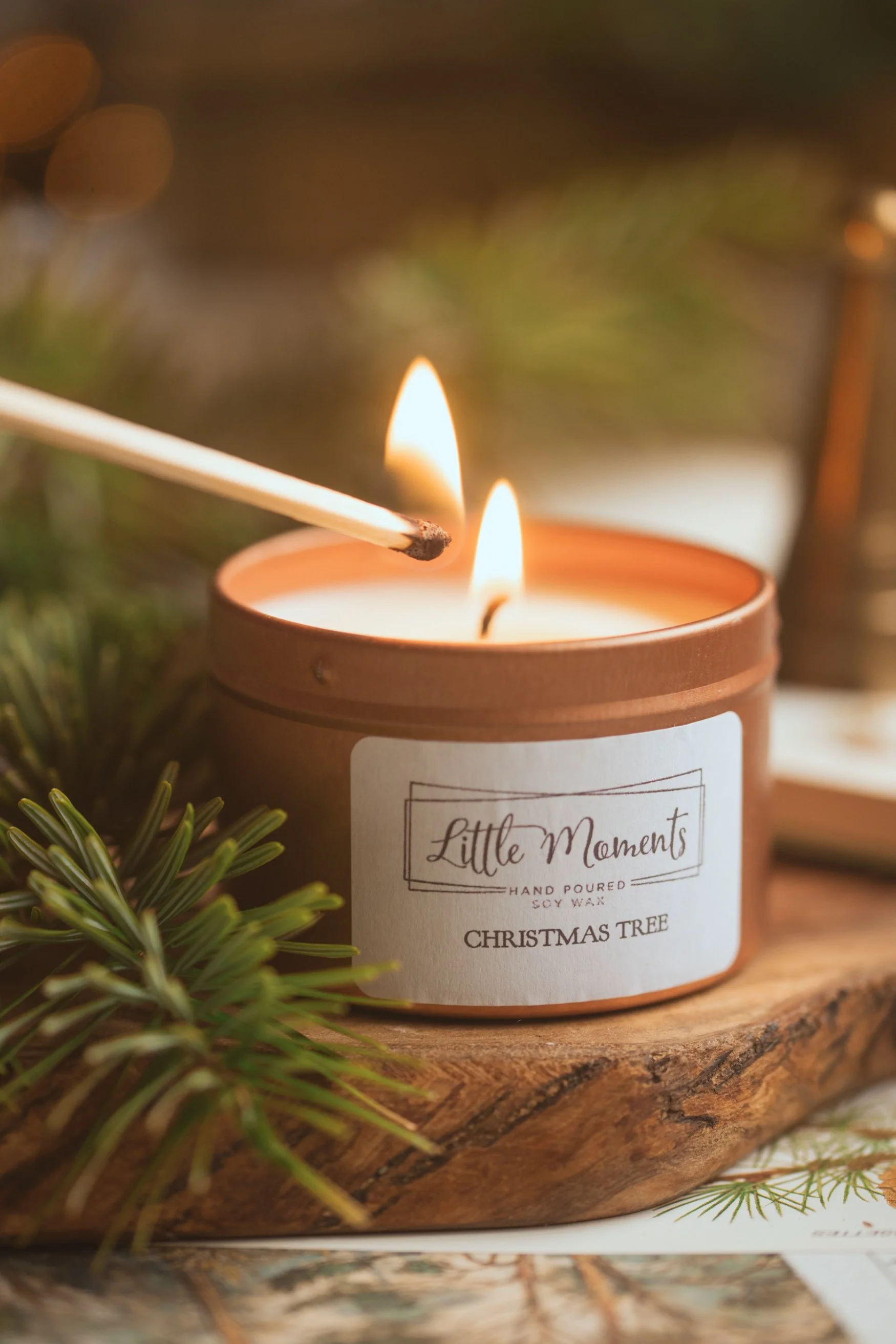A scented candle in a tin being lit with a match next to a sprig of fir