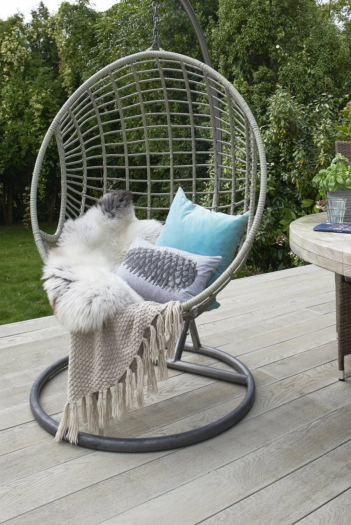 a freestanding egg swing chair styled with two cushions, a sheepskin and a hassled throw in a garden on a decking