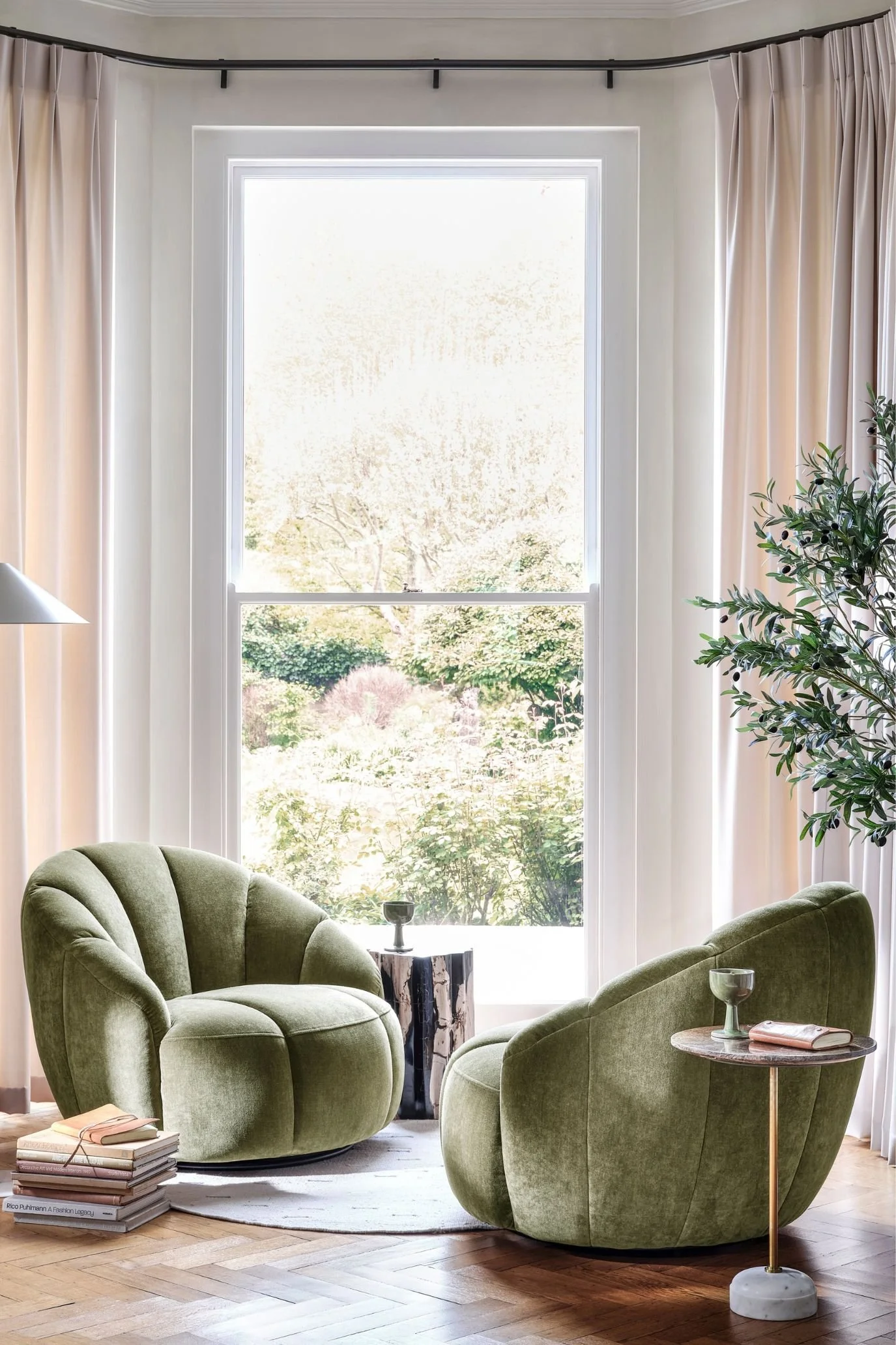 Two soft green curved swivel chairs beside a tall window framed by pale curtains, with small side tables.