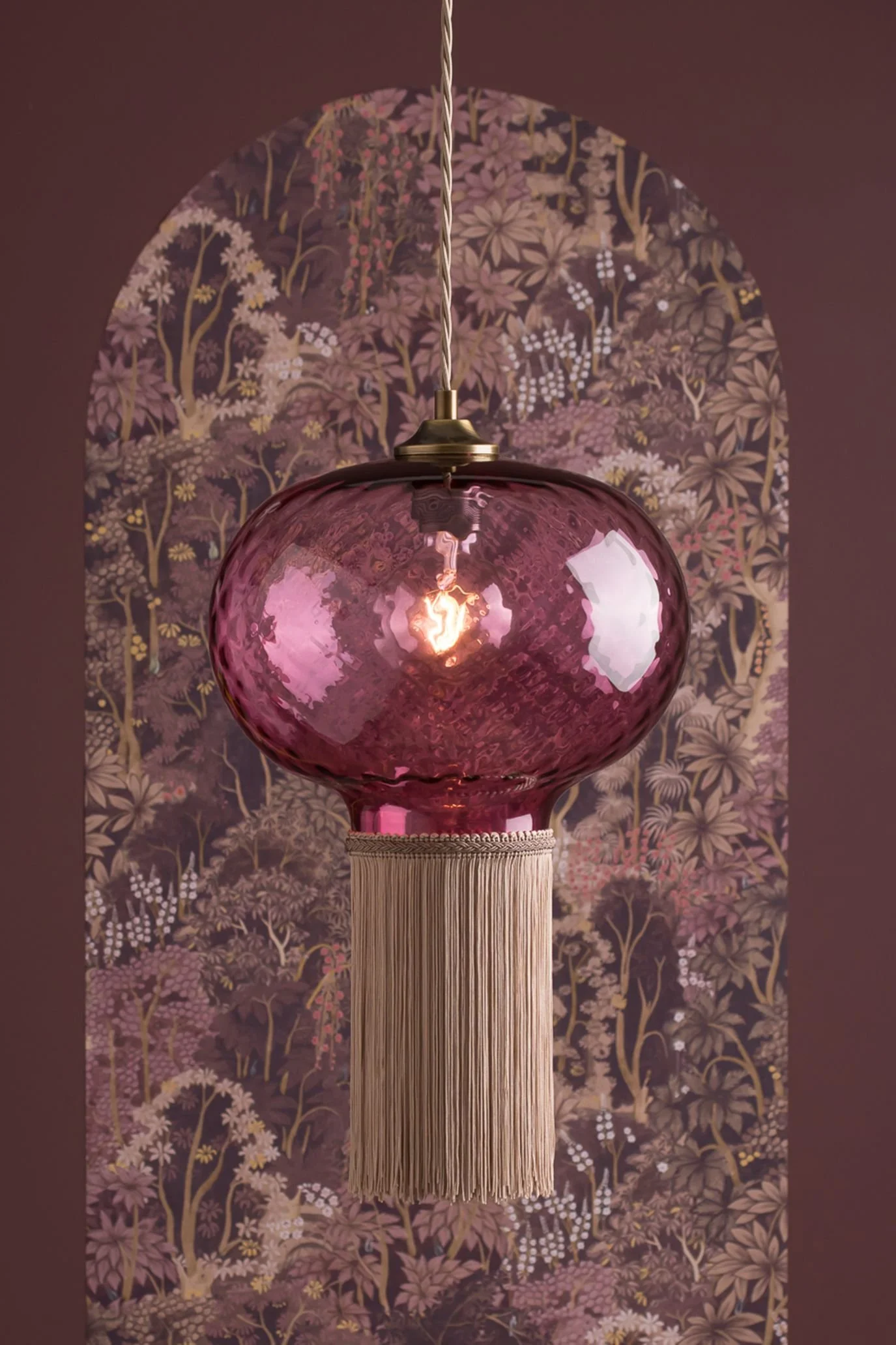 Pink hand-blown glass pendant light with a long decorative fringe against a patterned backdrop