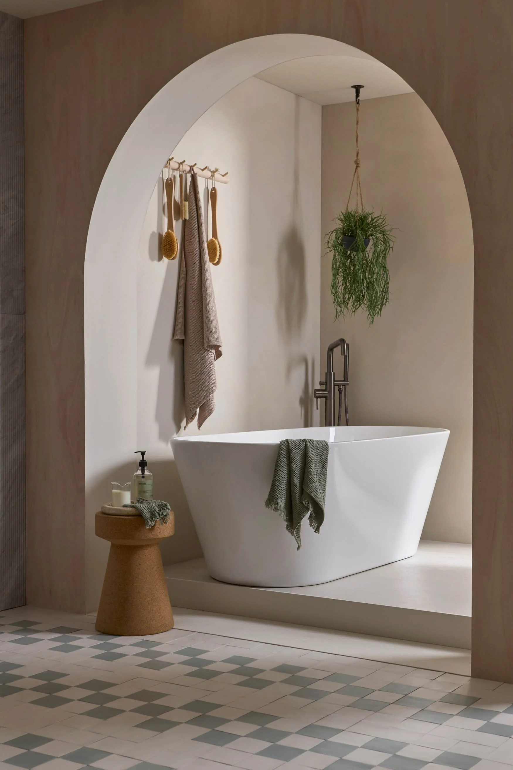 A freestanding bath and bronze bath filler behind an arch shaped wall with a hanging plant and side table