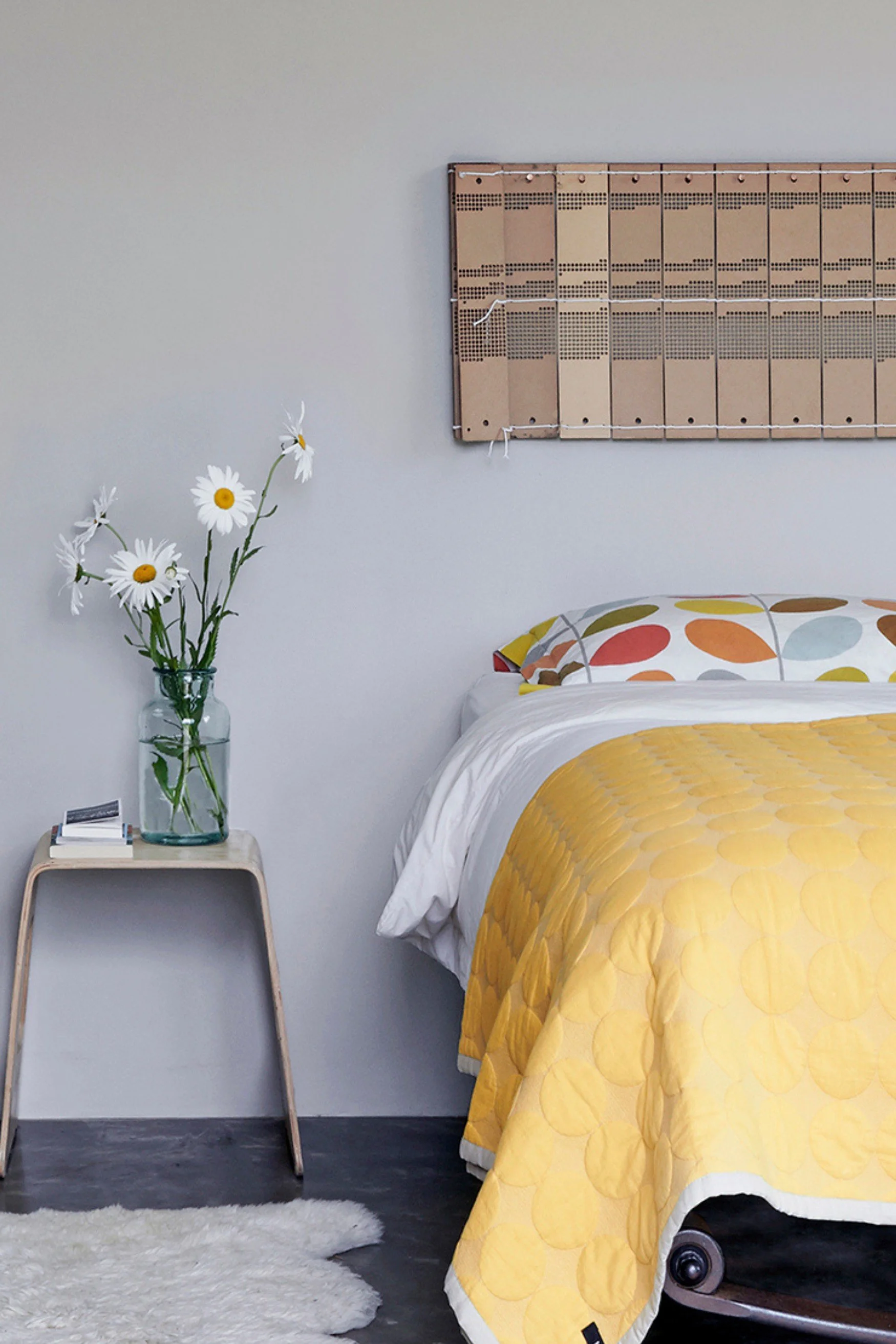 A bedroom with a bed with a yellow bedspread and patterned pillow cases in yellow and orange next to a bedside table with large white daisies in a vase. There is a headboard made out of a piano music roll
