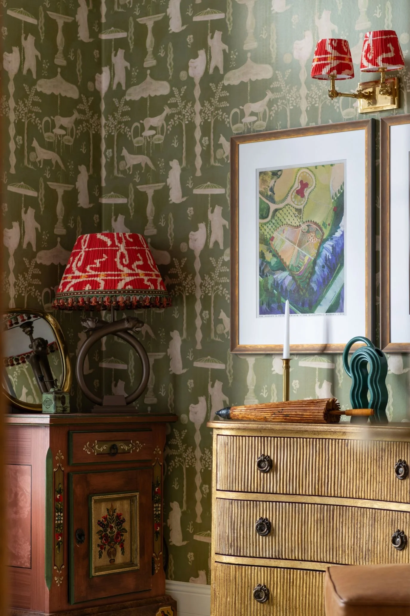 Framed green-toned artwork above a wooden chest of drawers against patterned olive green wallpaper.