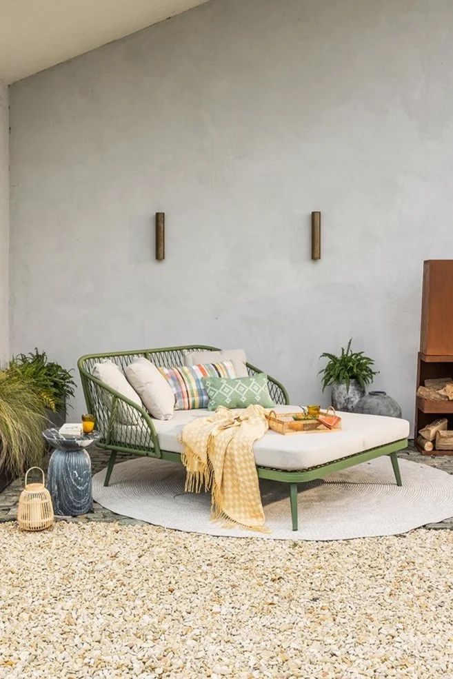 a green woven daybed, cushions and a throw, surrounded by plants, and a modern concrete wall with two outdoor lights.