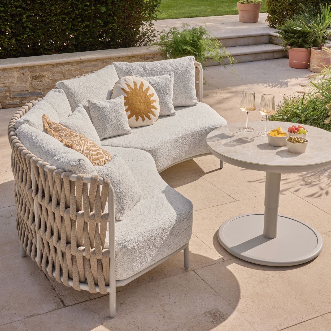 A curved outdoor sofa with boucle style cushions, a round sun cushion and another yellow-toned square cushion on a terrace next to a table
