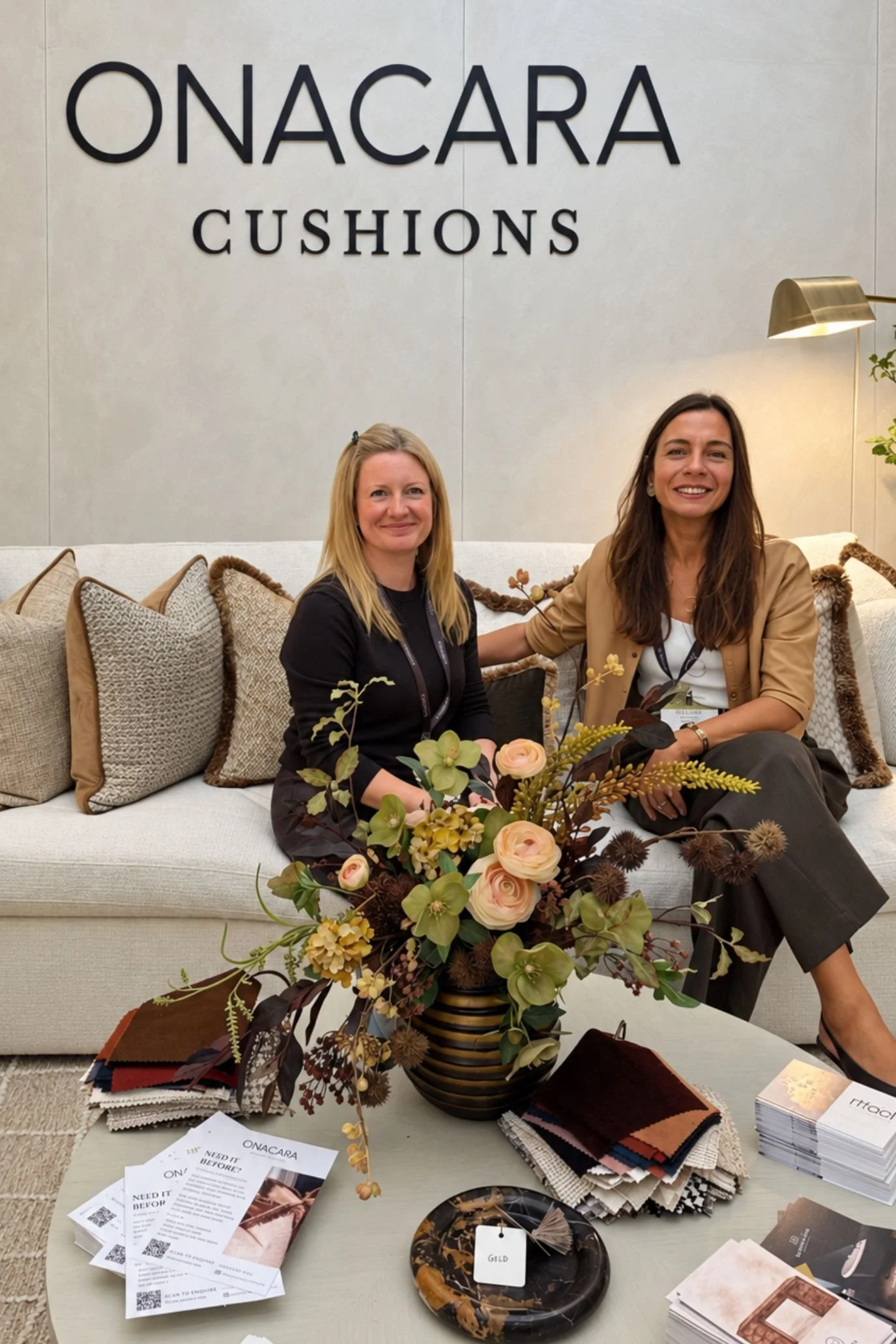 Two women seated on the Onacara stand, surrounded by cushions with decorative trims and a floral coffee-table arrangement.