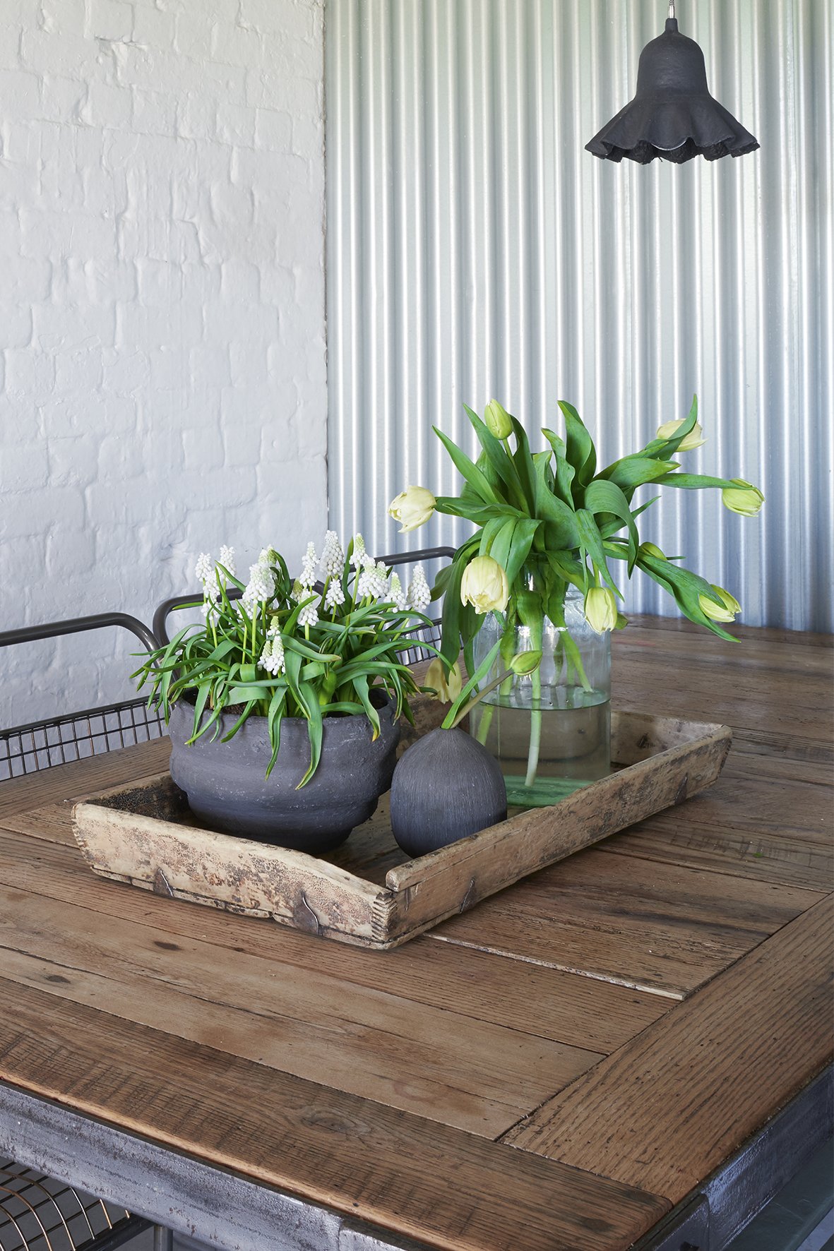 a wooden tray on a wooden table with a grey pot of white grape hyacinths and a vase of tulips with a corrugated iron wall and black pendant light