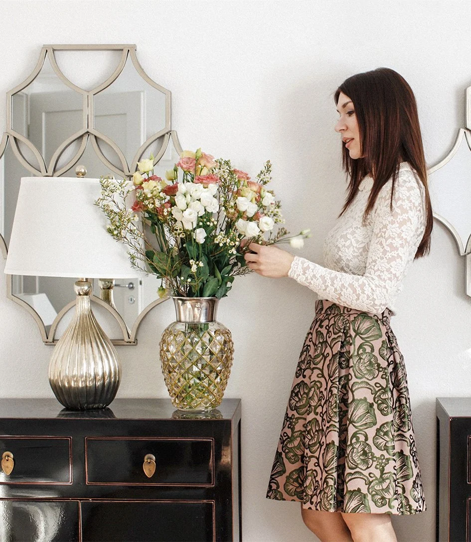 Interior Designer Katie Fischer arranging a large display of flowers in a glass vase next to a silver based lamp with white shade