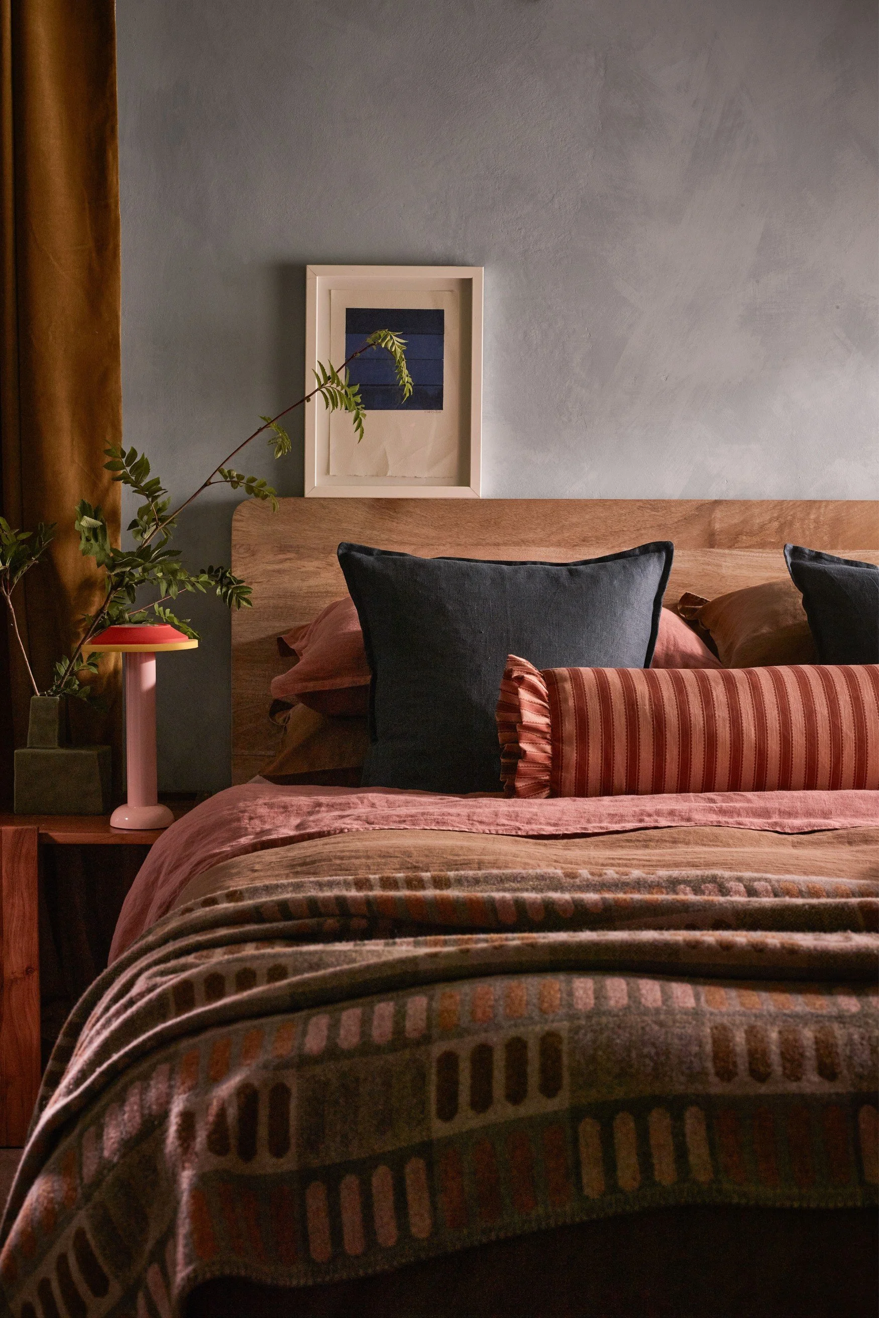 A bed dressed in terracotta tones with a printed throw, striped bolster cushion and a mango wood headboard.