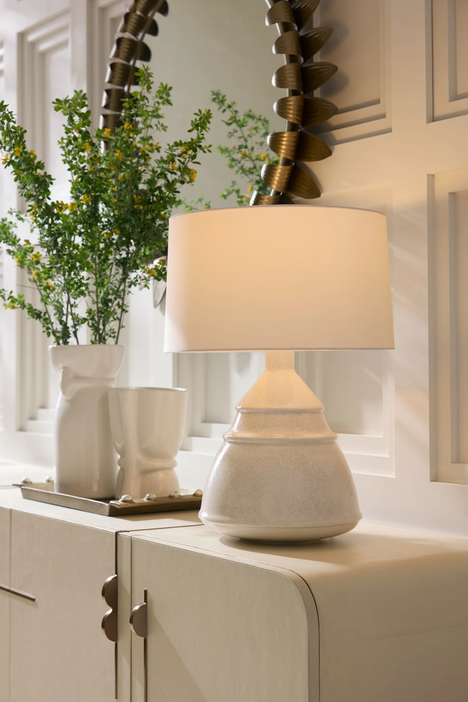 A lamp and two vases in off-white on a sideboard with greenery and a bronze circular mirror