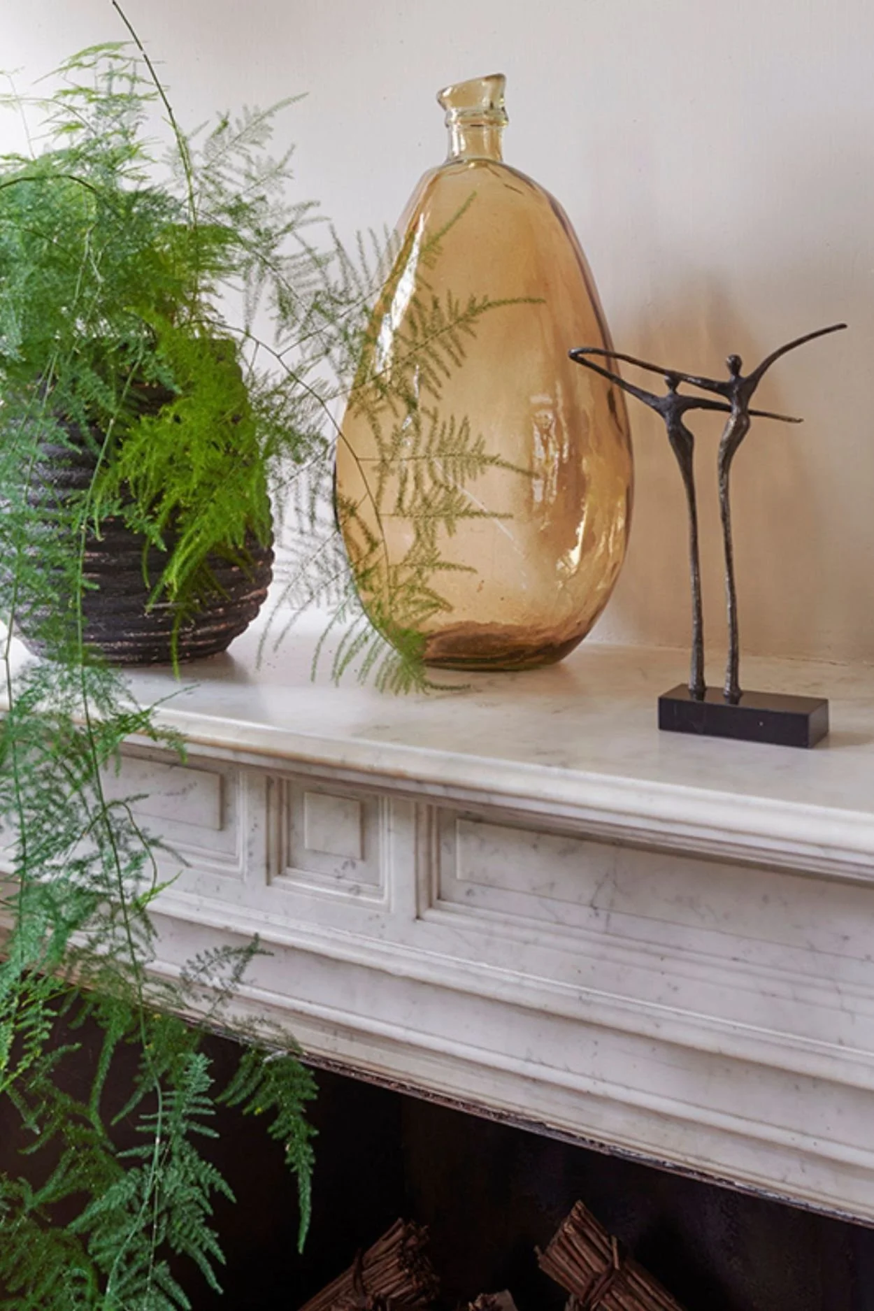 Stylish home decor vase in amber glass, an asparagus fern in a black ribbed pot and two dancing figurines on a marble mantle.
