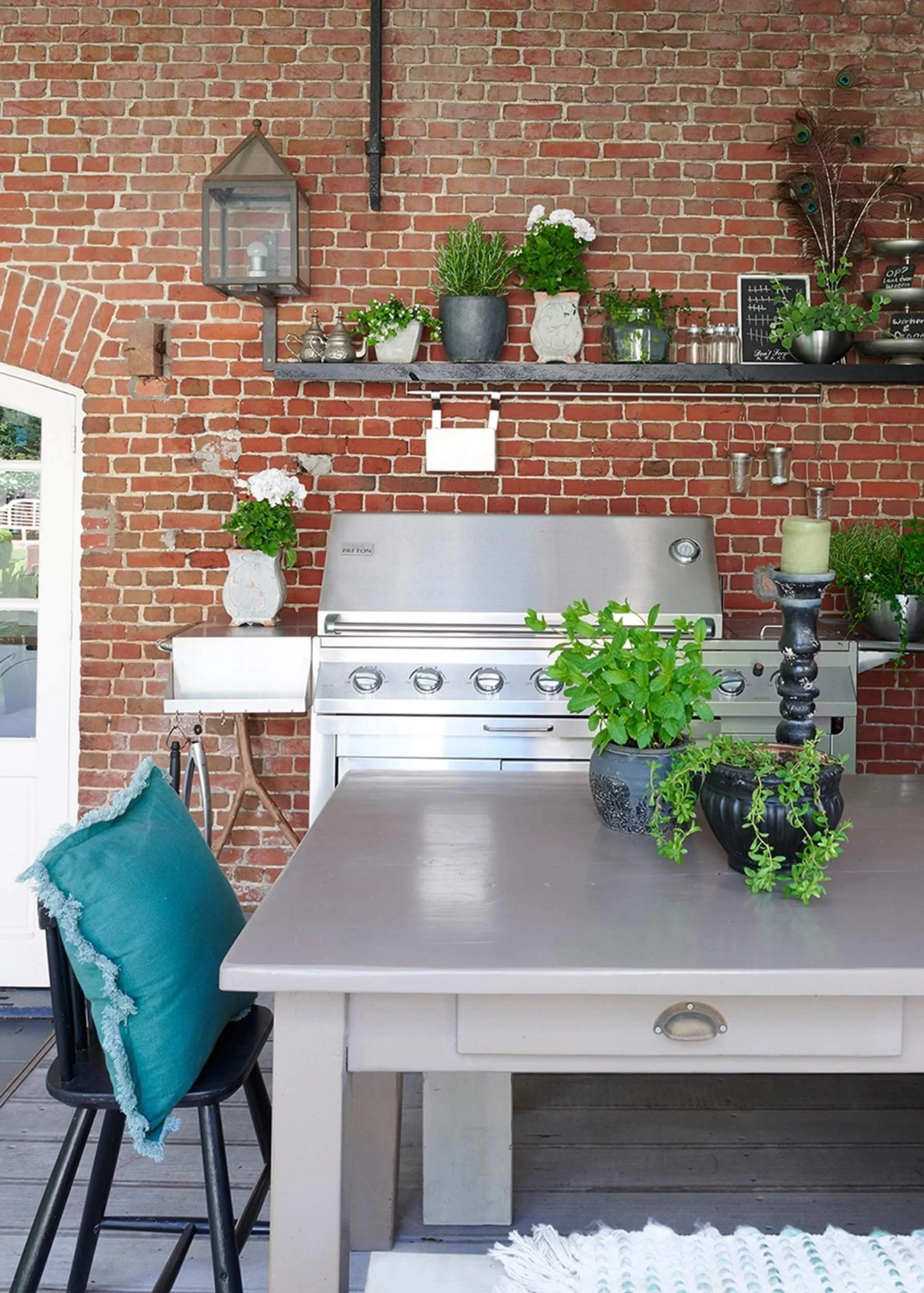 outdoor kitchen with large range bbq, table and shelving with a selection of potted plants and herbs set against a brick wool in an outdoor undercover area