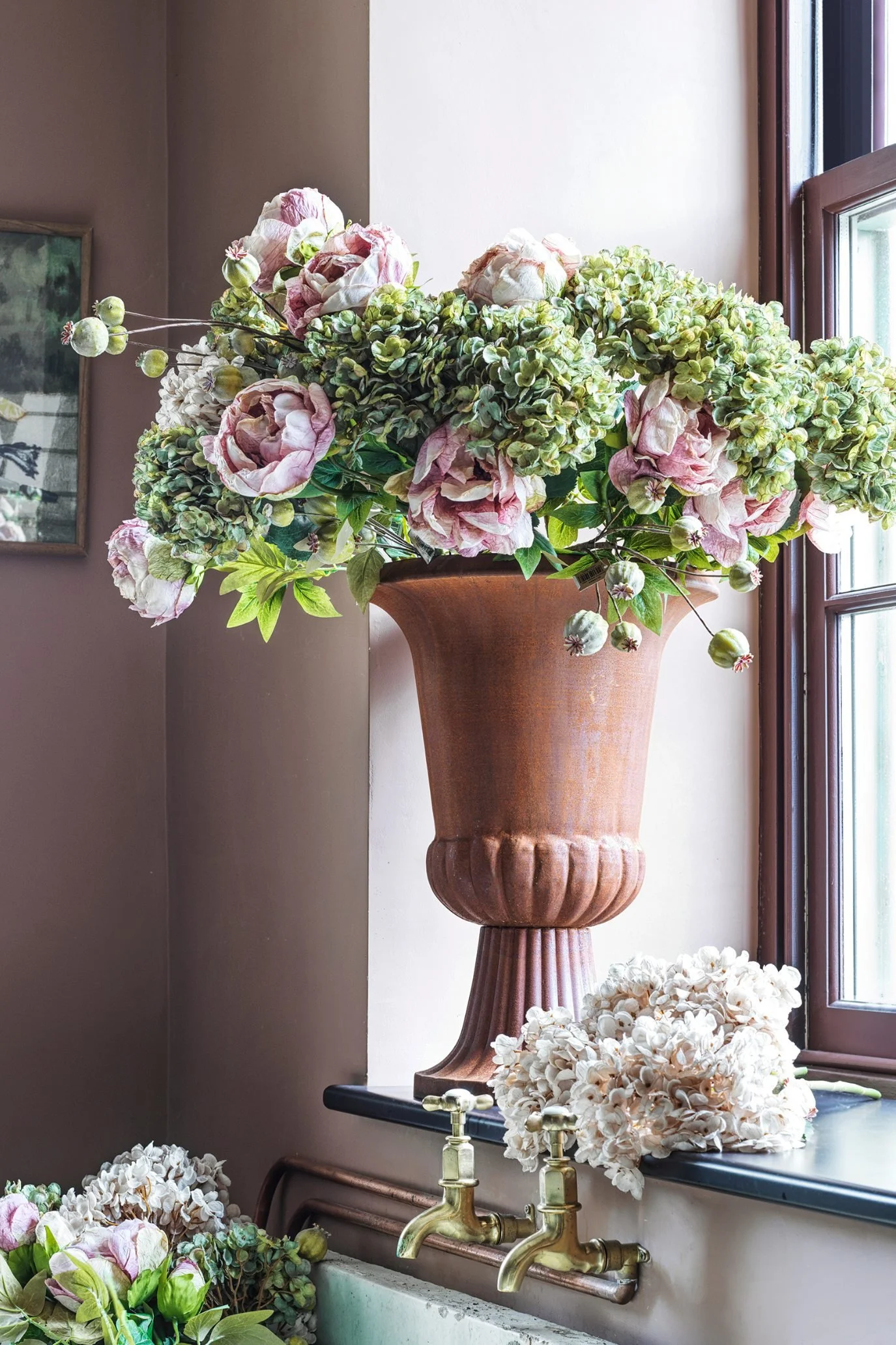 Faux peonies and green hydrangeas arranged in a large terracotta urn by a window.