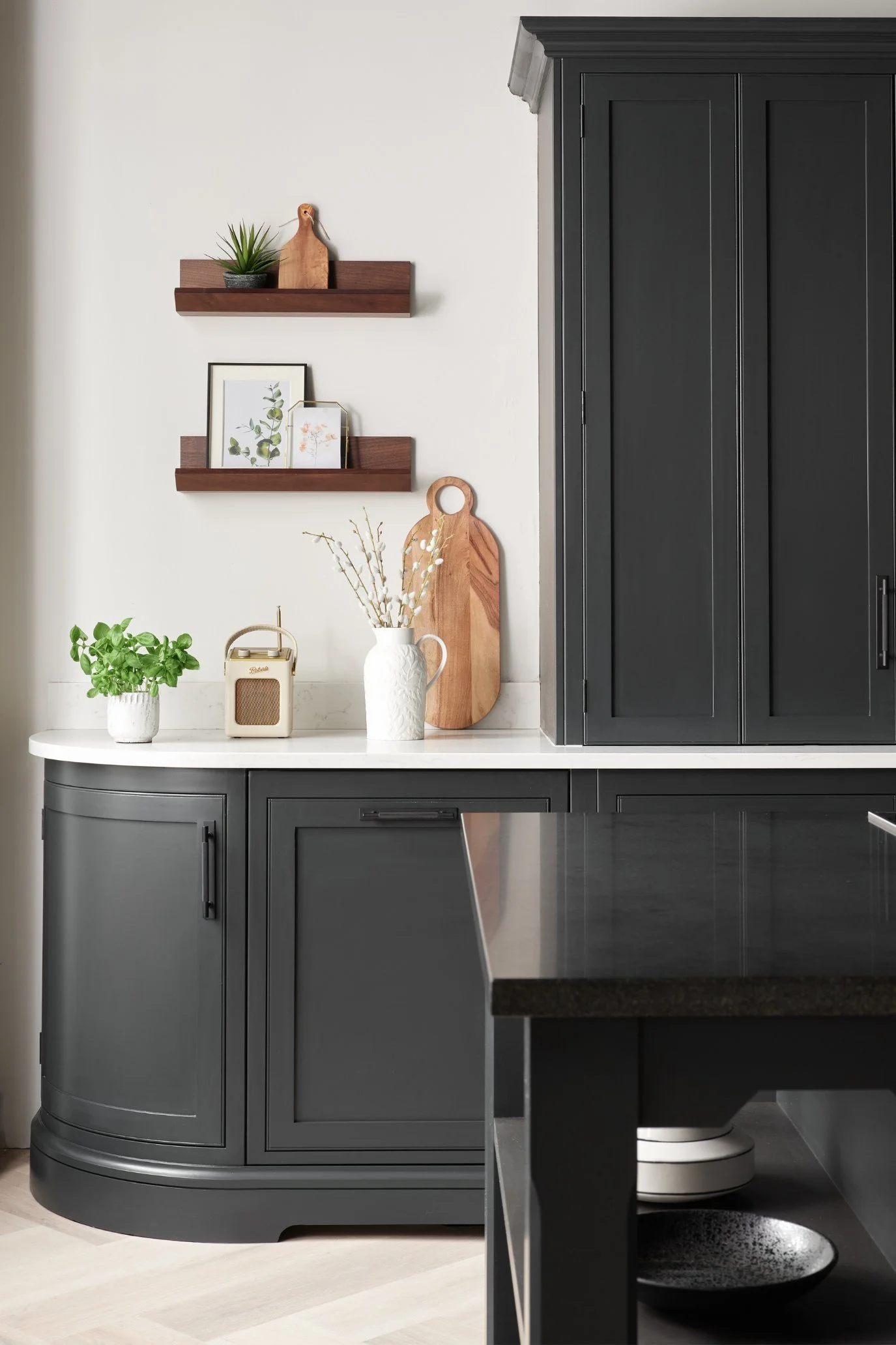 A contempoary dark grey kitchen with a white countertop decorated with plants, a wooden chopping board and a radio. There a two dark wood picture ledges with pictures on the bottom and a chopping board and plant on the top shelf