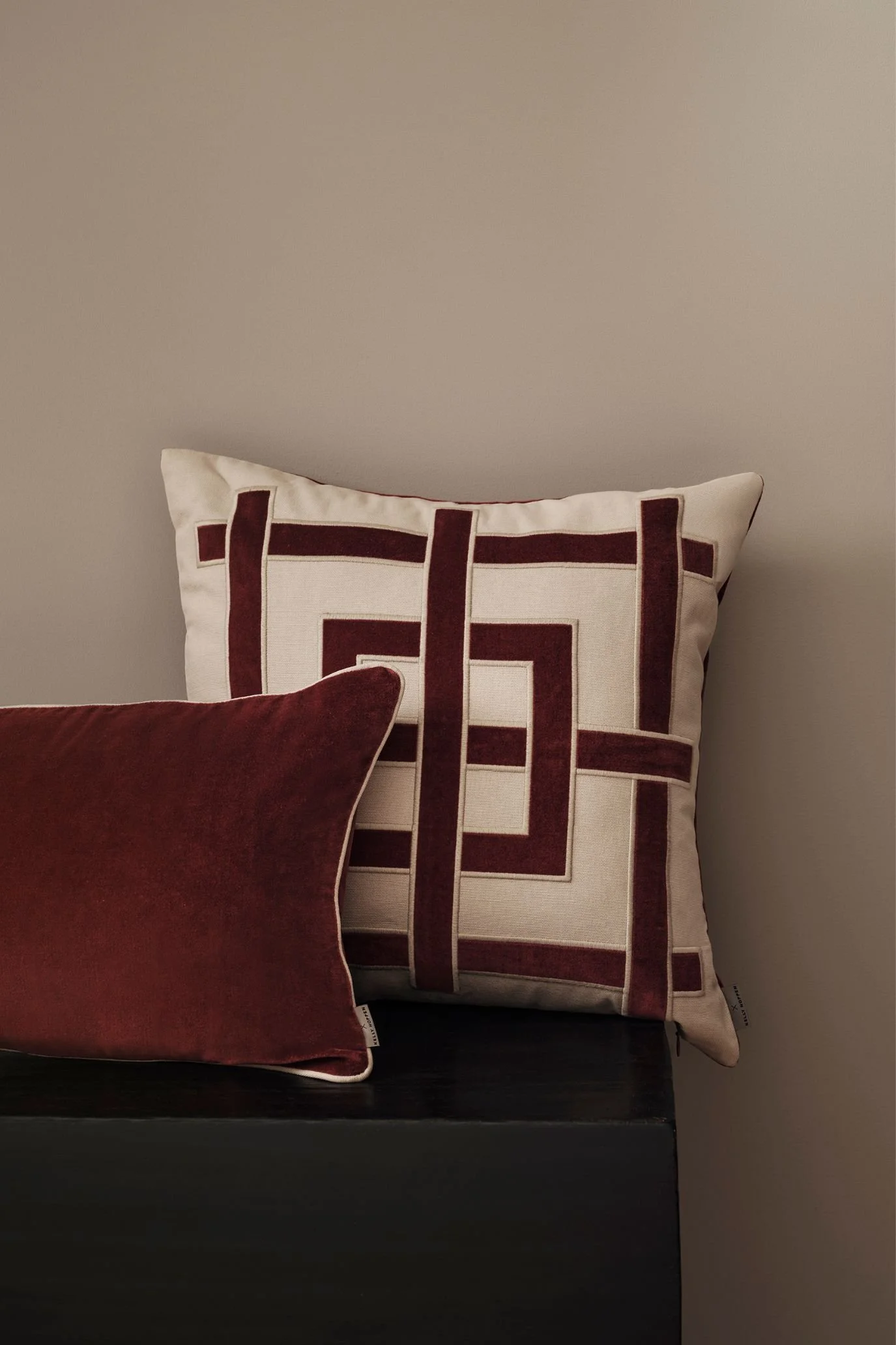 Kelly Hoppen x M&S cream cushion with burgundy geometric detailing, paired with a plain burgundy cushion on a dark sofa.
