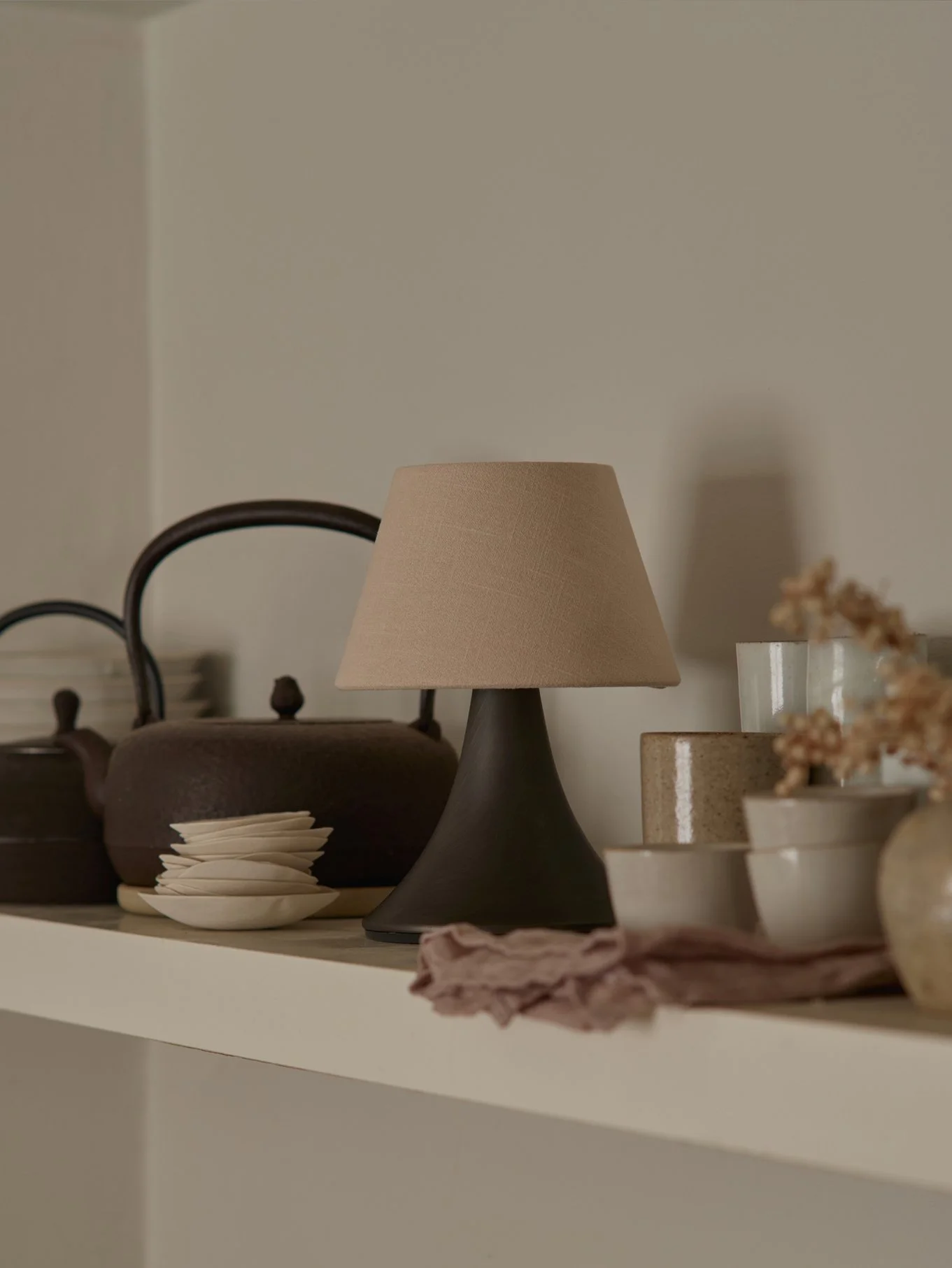 A bronze and linen lamp next to two vintage tea pots and stacked earthenware cups