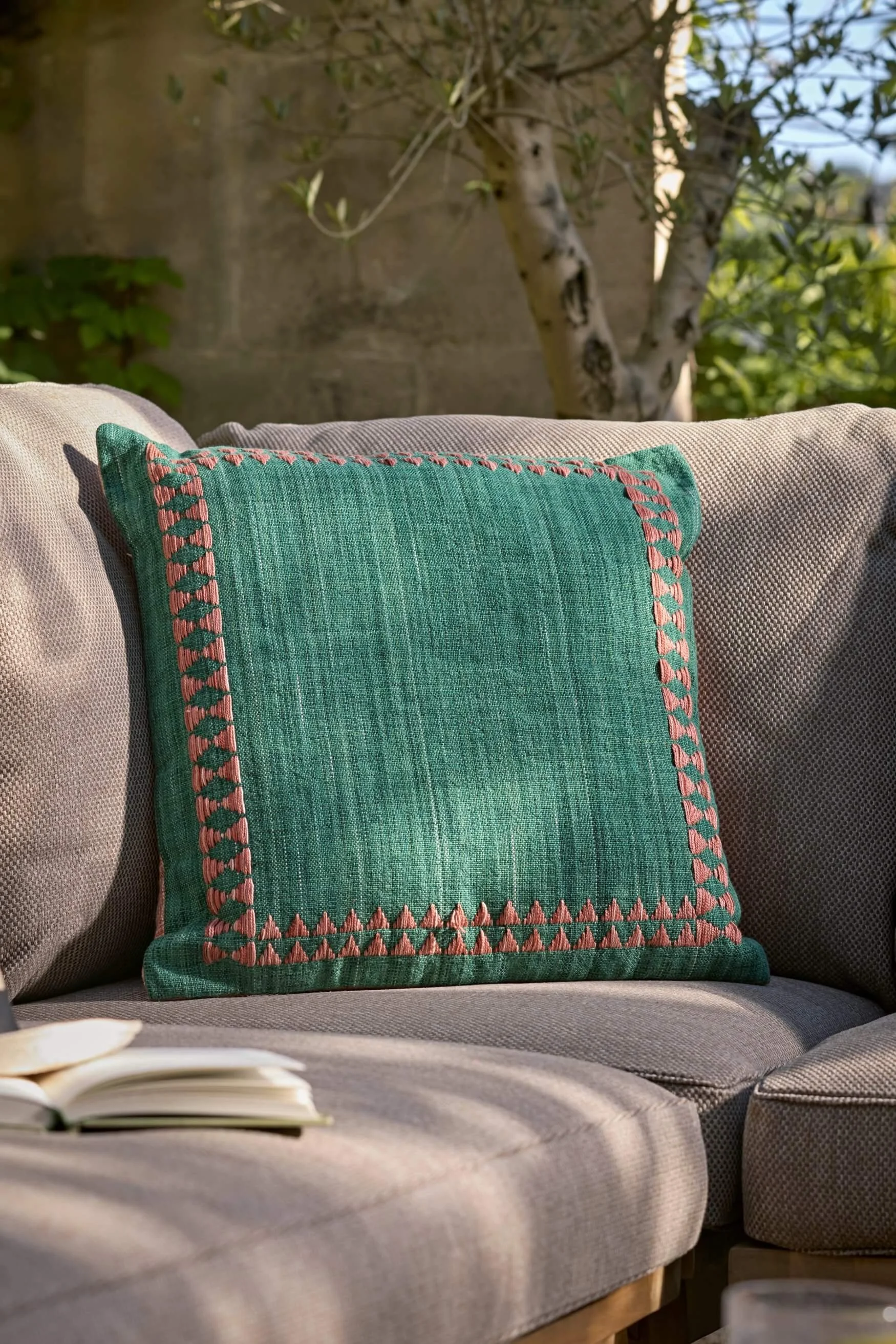 Square green outdoor cushion with embroidered terracotta trim on a beige sofa