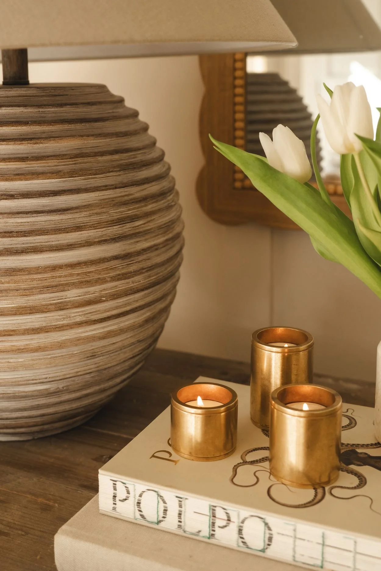 Three tea lights in brass holders on top of books and next to tulips and a ribbed wooden lamp