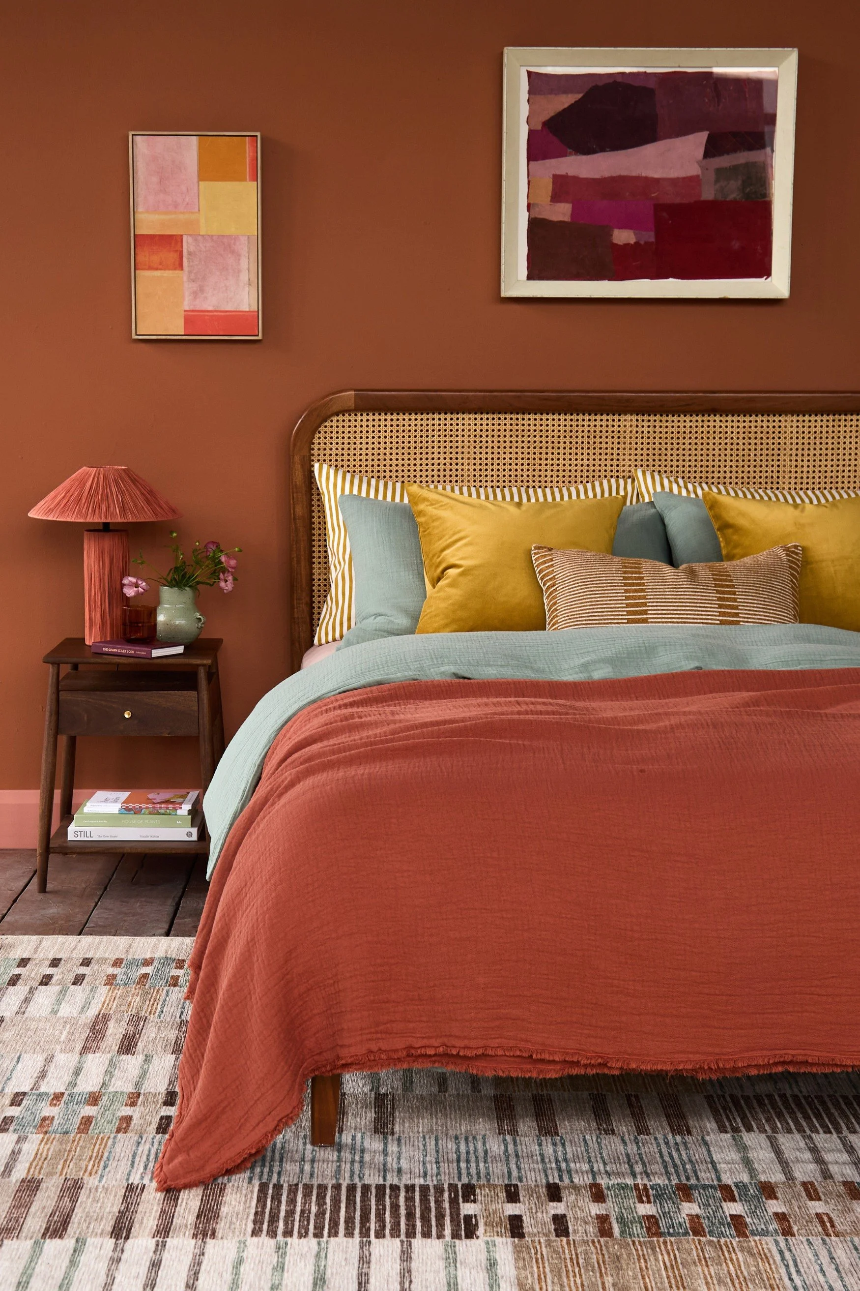 A rattan and dark wood bed dressed with blue and gold cushions and pillows and a terracotta throw that pairs with the wall in the same tone