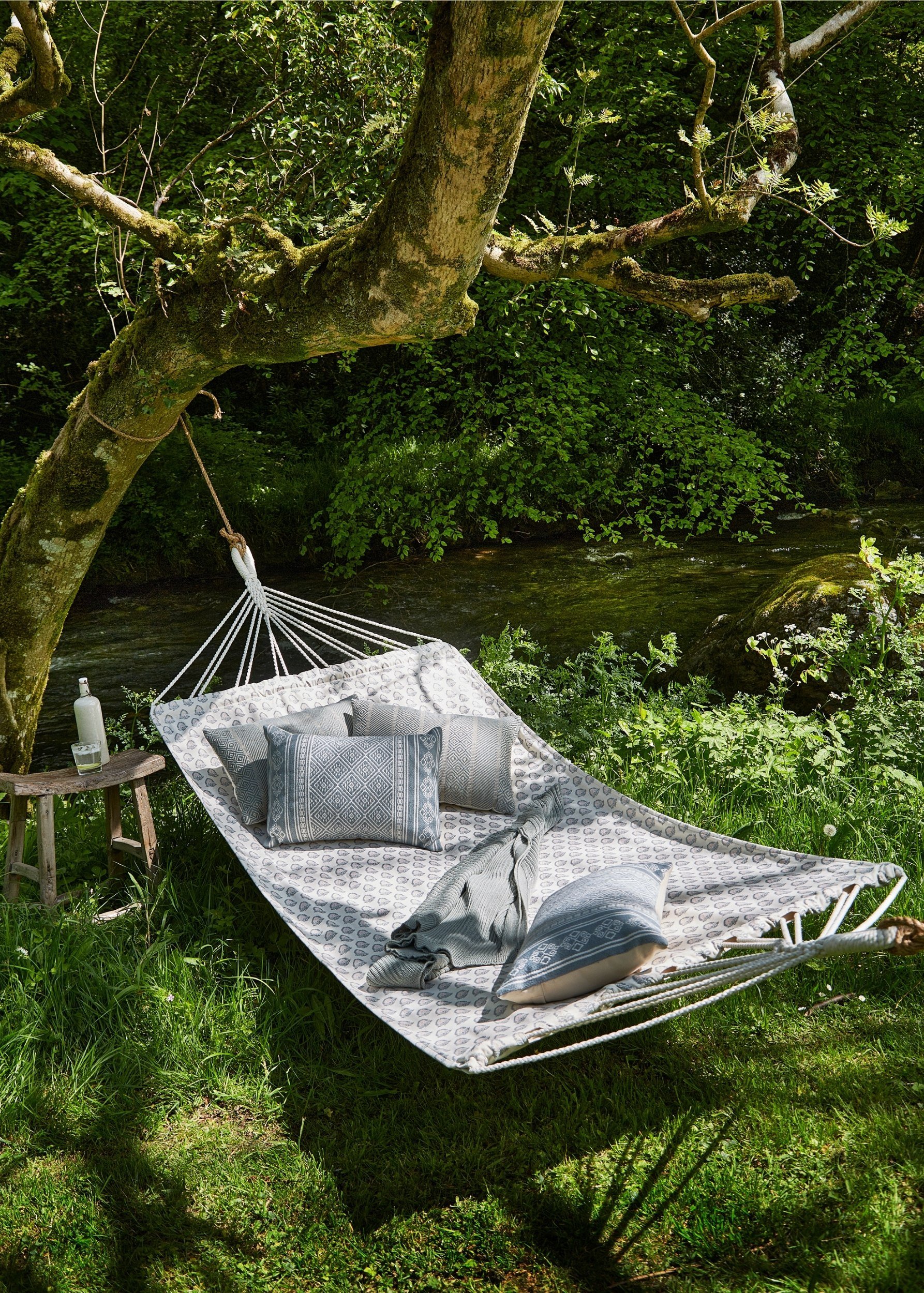 A large outdoor printed hammock with cushions on it tied to a tree next to a river