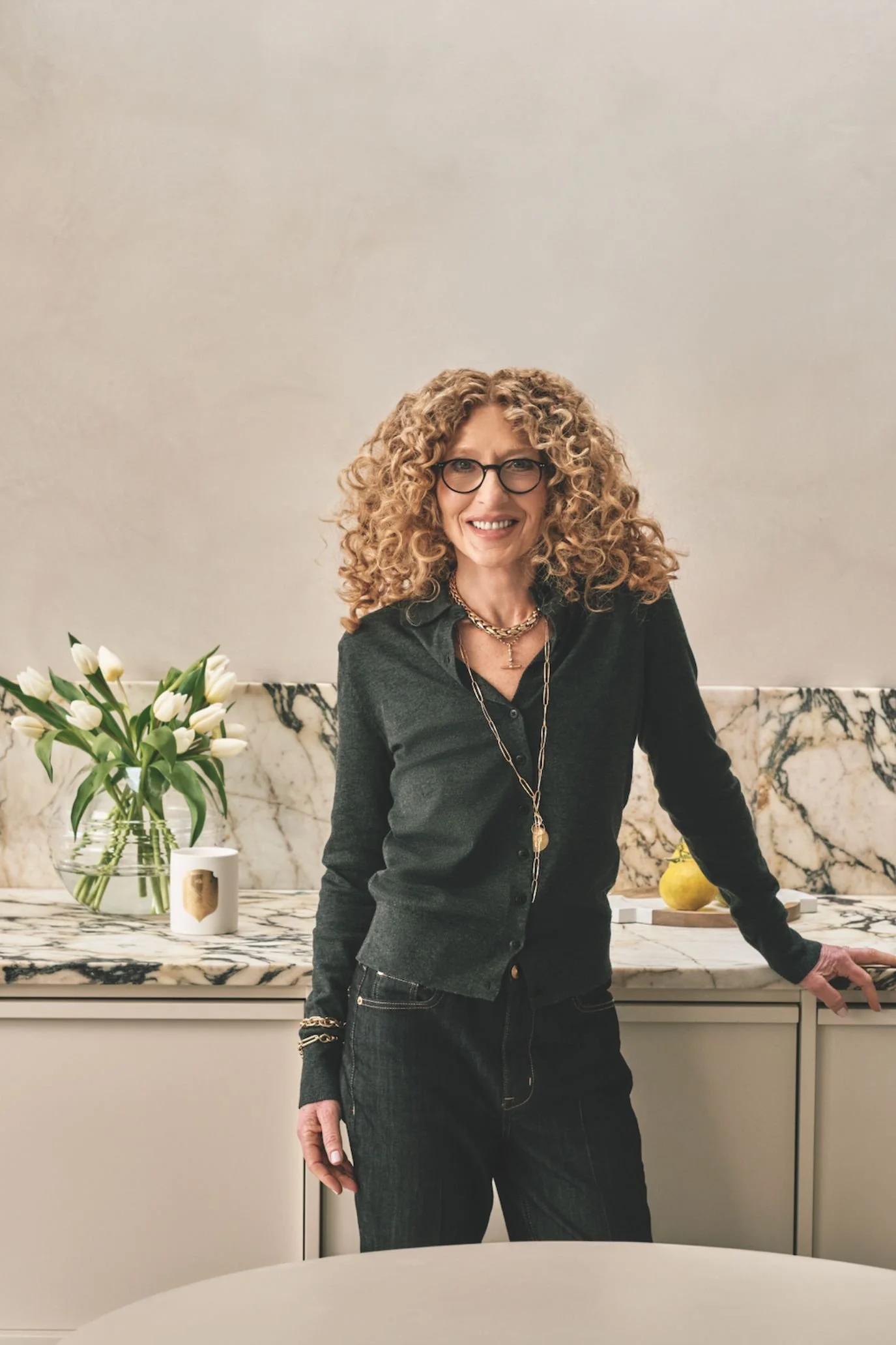 Kelly Hoppen wearing a dark jacket, standing in a neutral kitchen with flowers and decorative accessories.