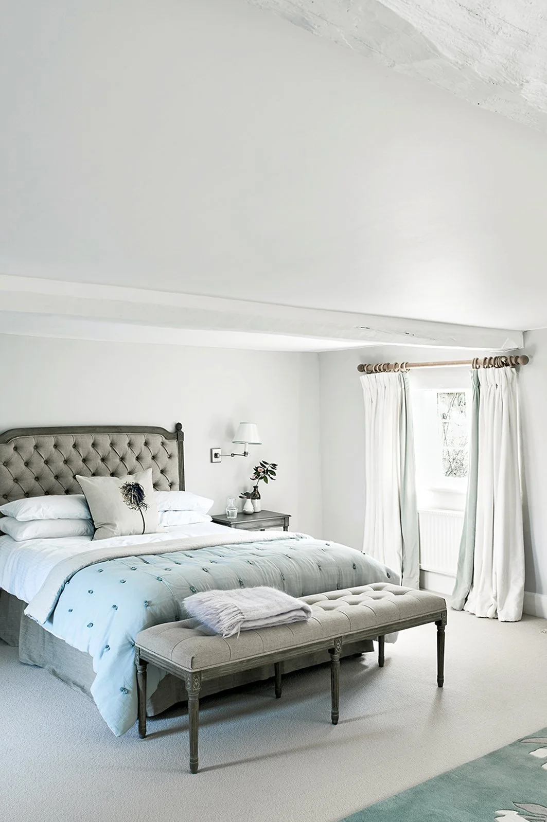 Bedroom in white with a beige upholstered bed and ottoman with a jade silk throw and a cushion with a flower