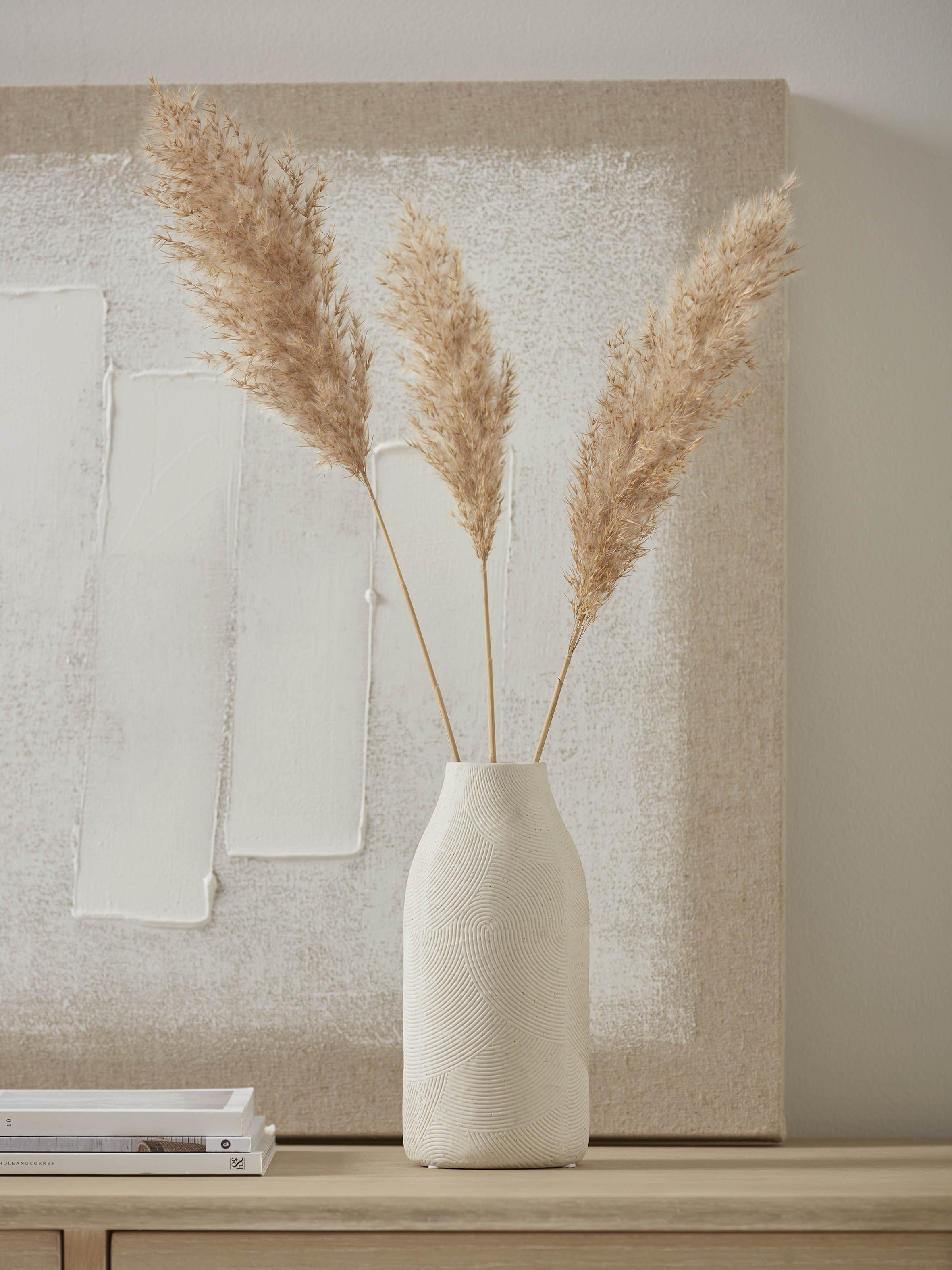 Three dried pampas grasses in a cream vase with a  painting