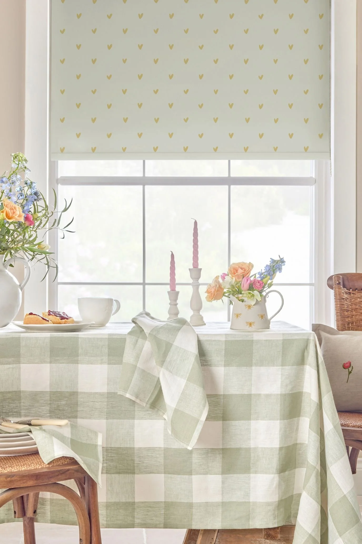 Dining table with a sage green checked tablecloth beneath a pale patterned roller blind, styled with flowers and candles.