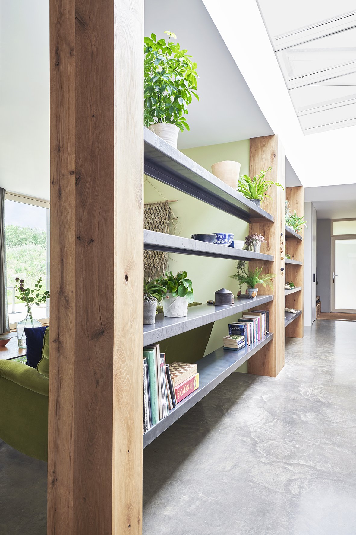 Shelving with zinc shelves and large oak posts runs the whole length of two rooms with plants and books on it.