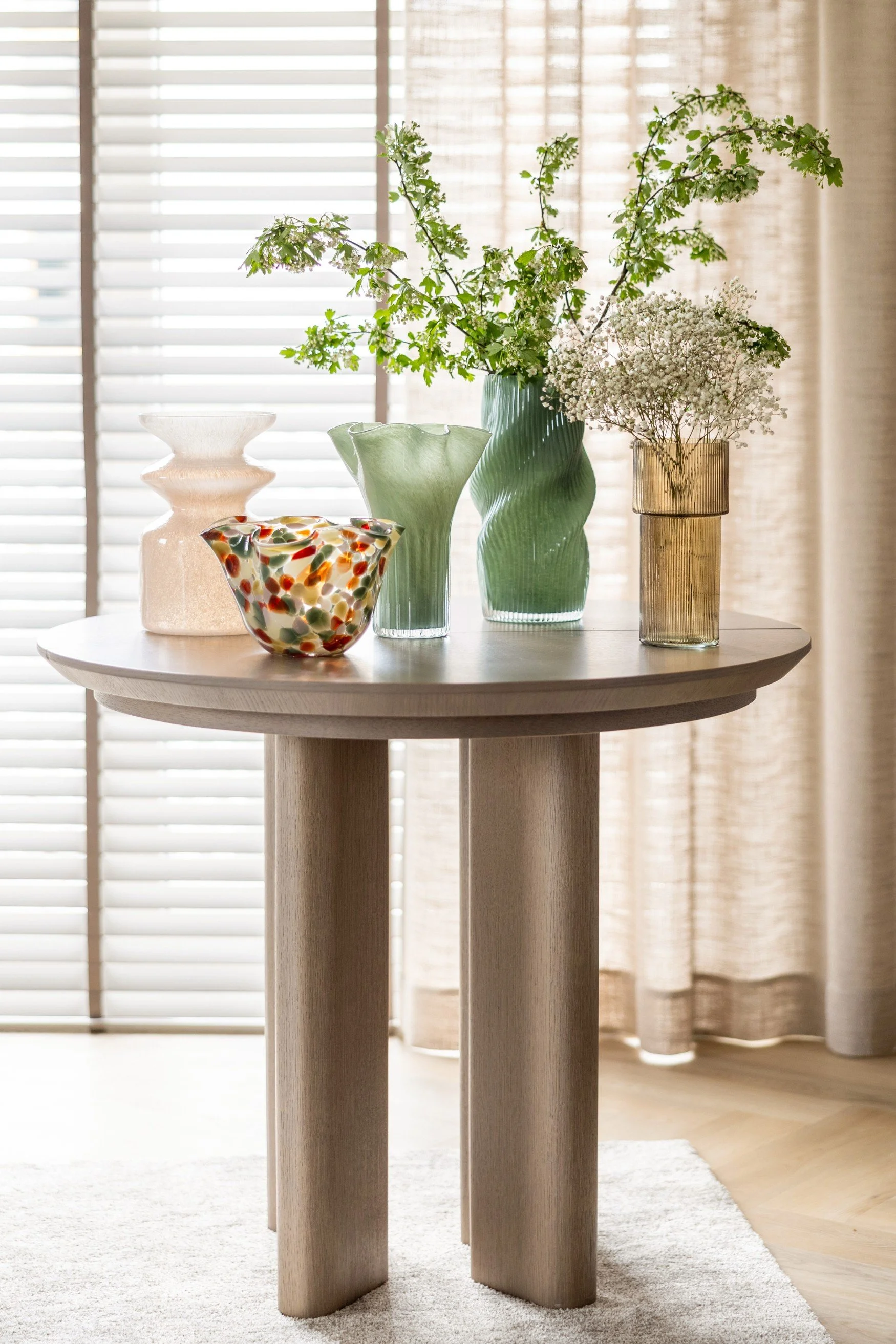 A side table with 5 different shaped vases, two soft green, one pink, one light brown and a multi coloured shorter one