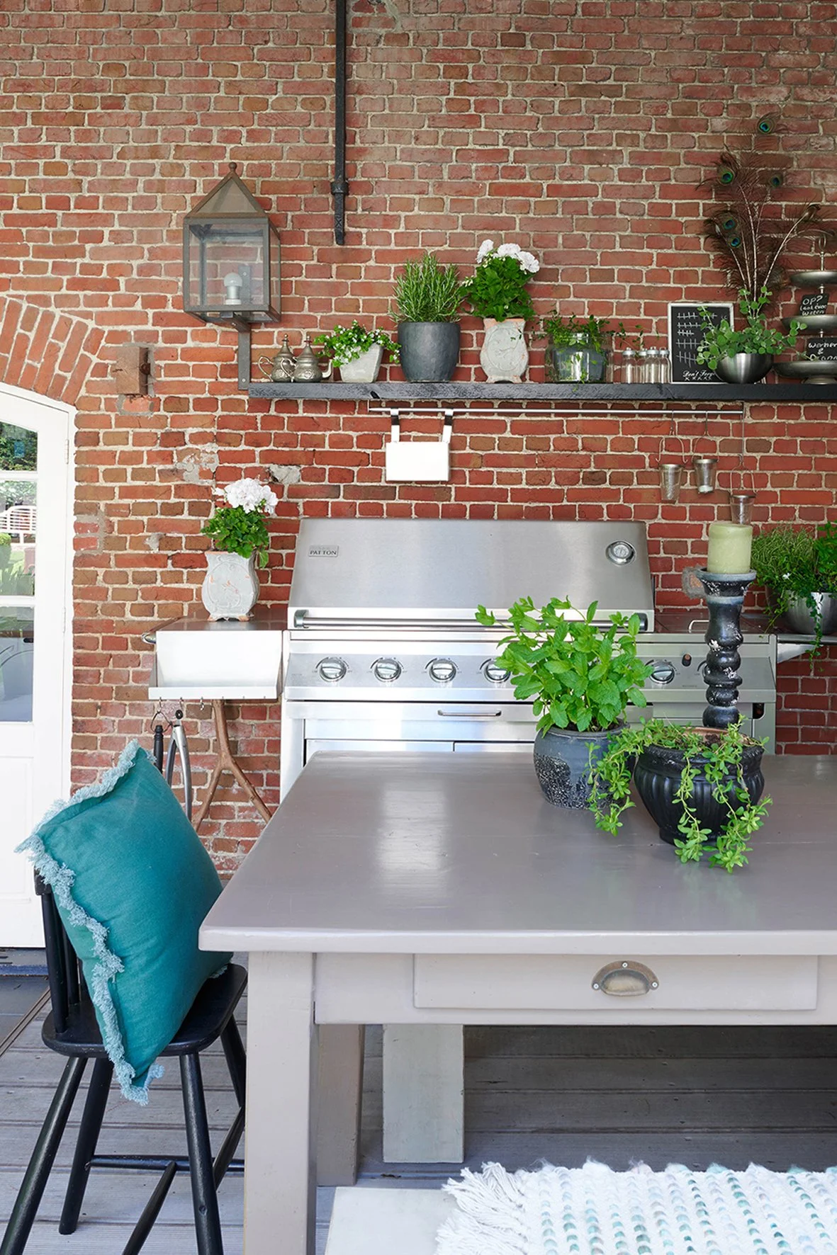outdoor kitchen with large range bbq, table and shelving with a selection of potted plants and herbs set against a brick wool in an outdoor undercover area