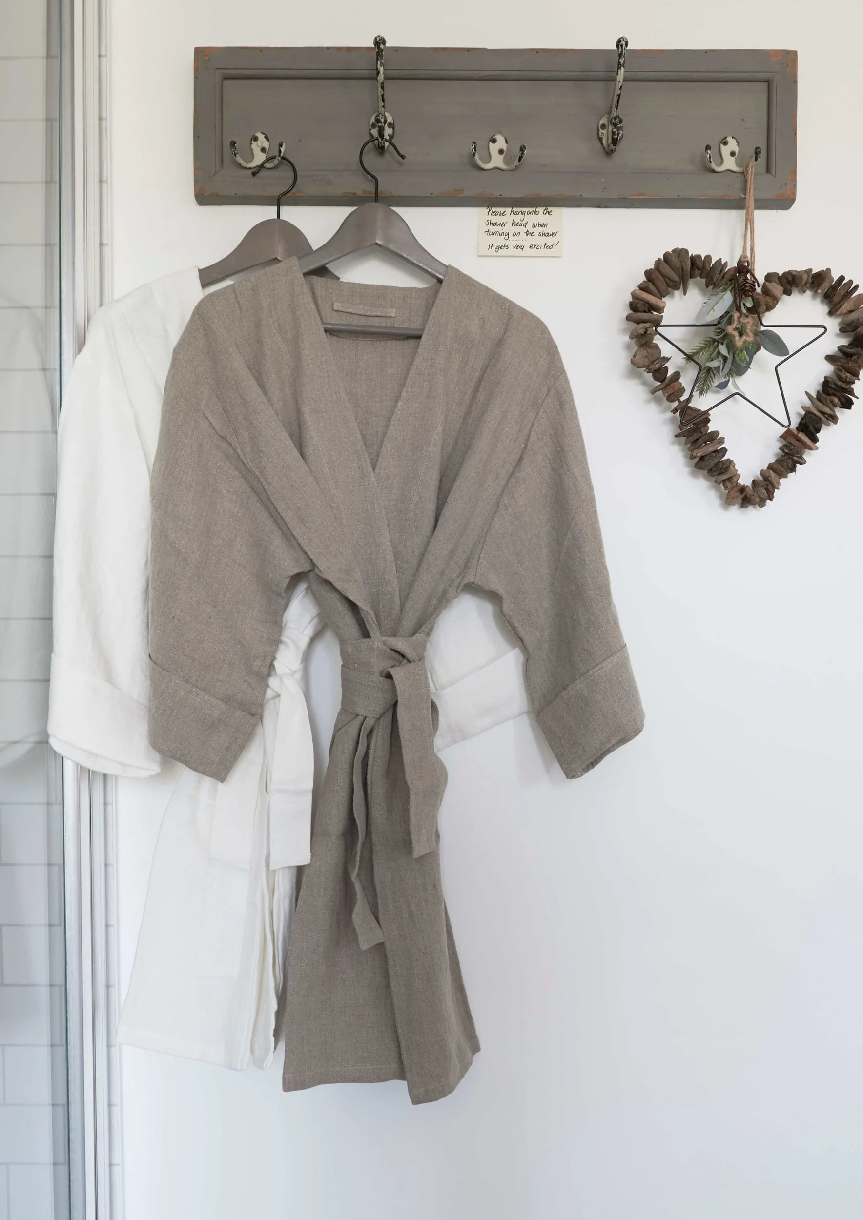 A beige and white robe hang from vintage hooks on a wooden plinth with a heard made of Pebles hanging next to it
