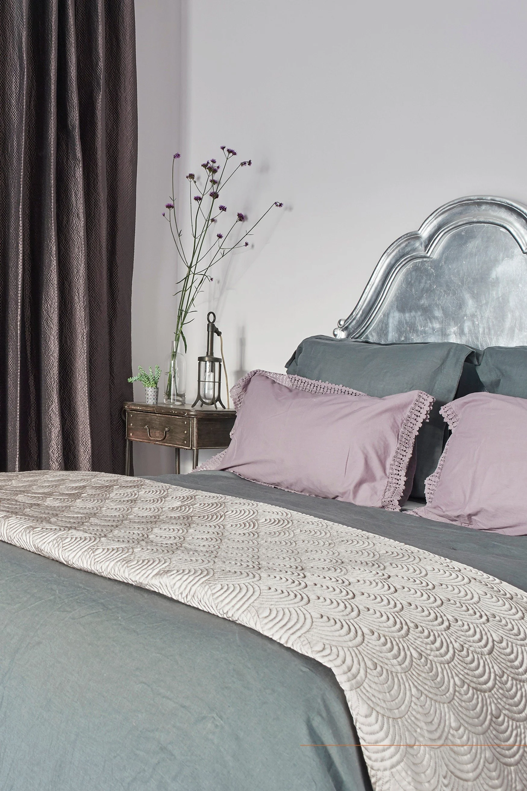A bedroom with a silver headboard and bed linen in grey and pink with a dark mauve silk curtain and flowers and a lamp on a metal side table