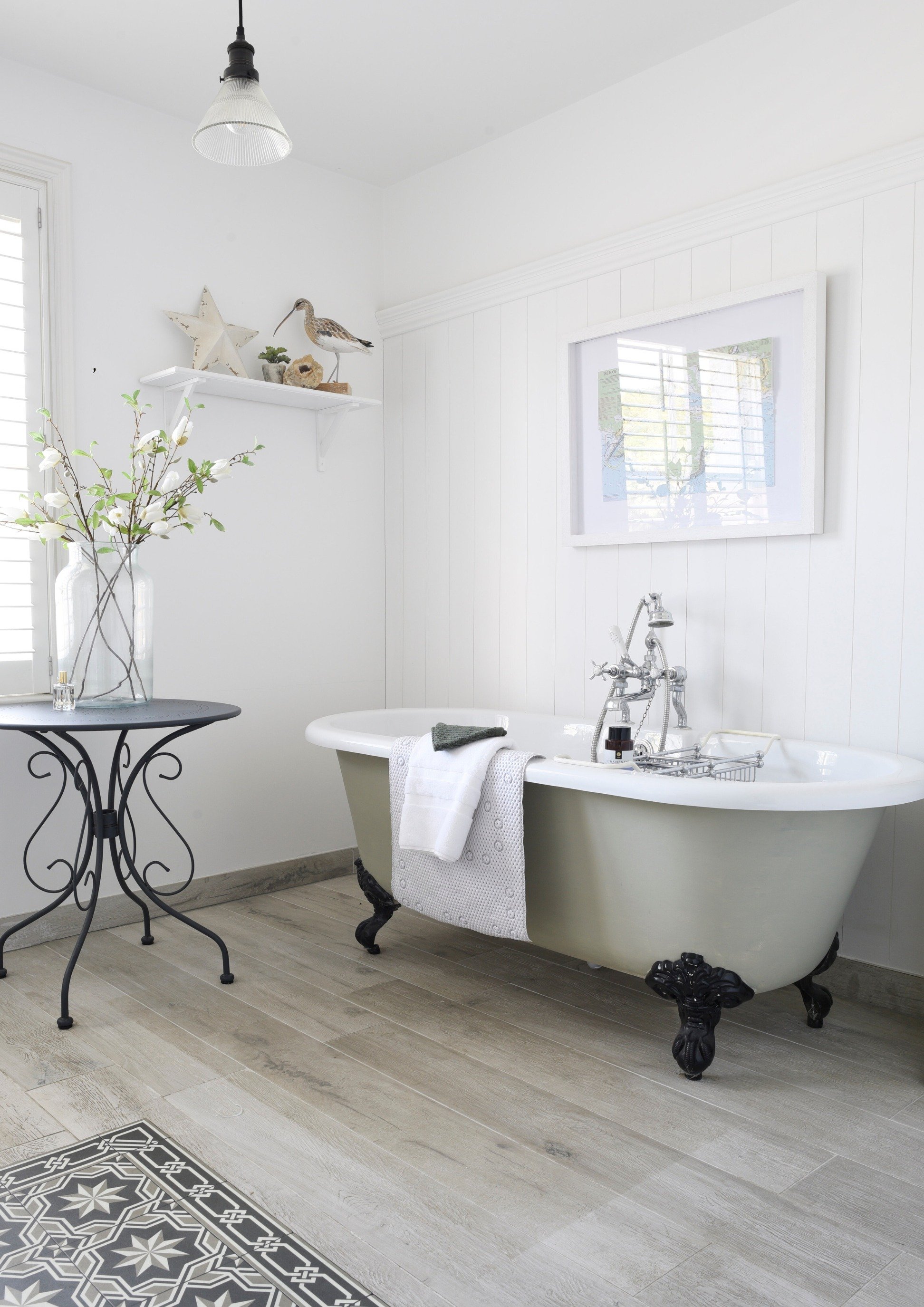 A soft green roll top bath sits on a wooden floor beneath a framed print and near a shelf with a statue of a sea gull. There is a table with a vintage vase and white flowers