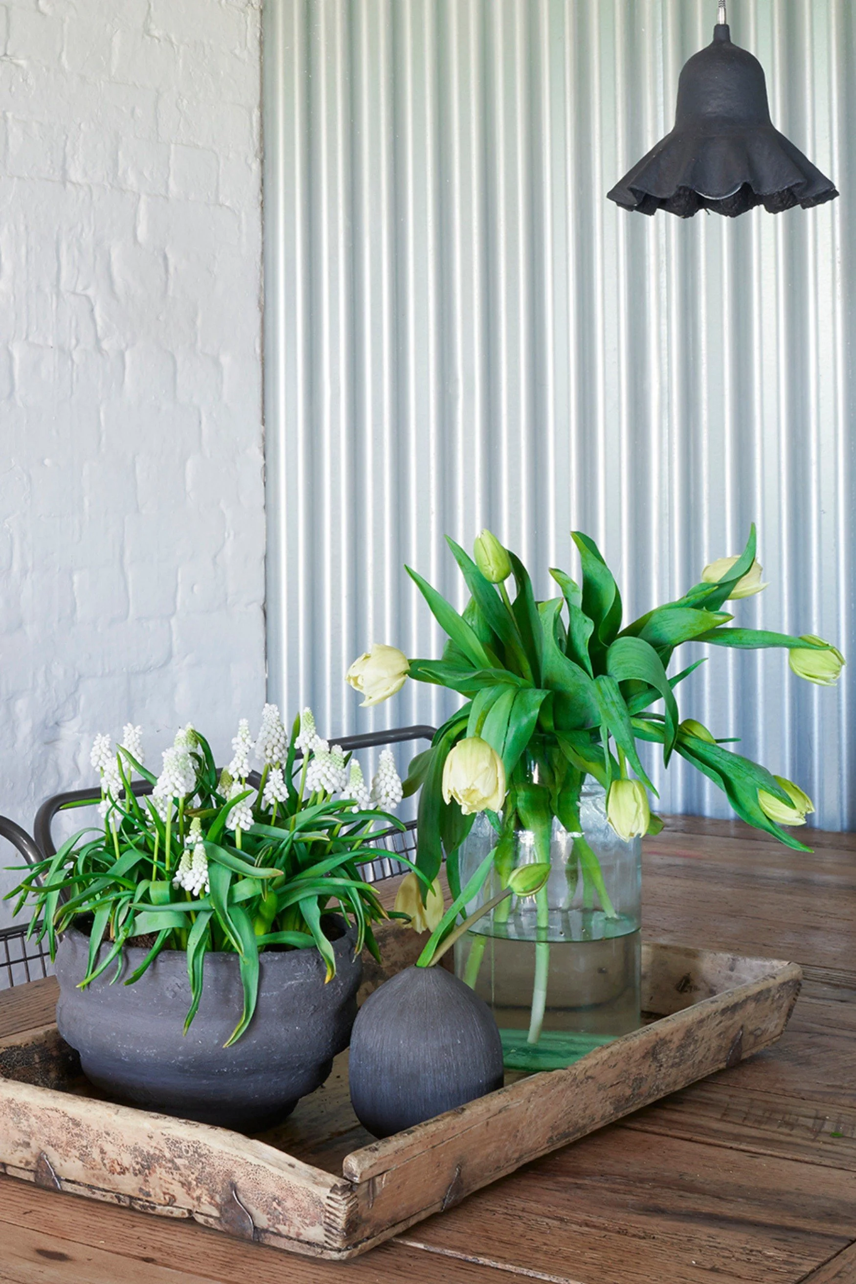 A wooden tray on a wood table with grey pots and a vase with spring flowers
