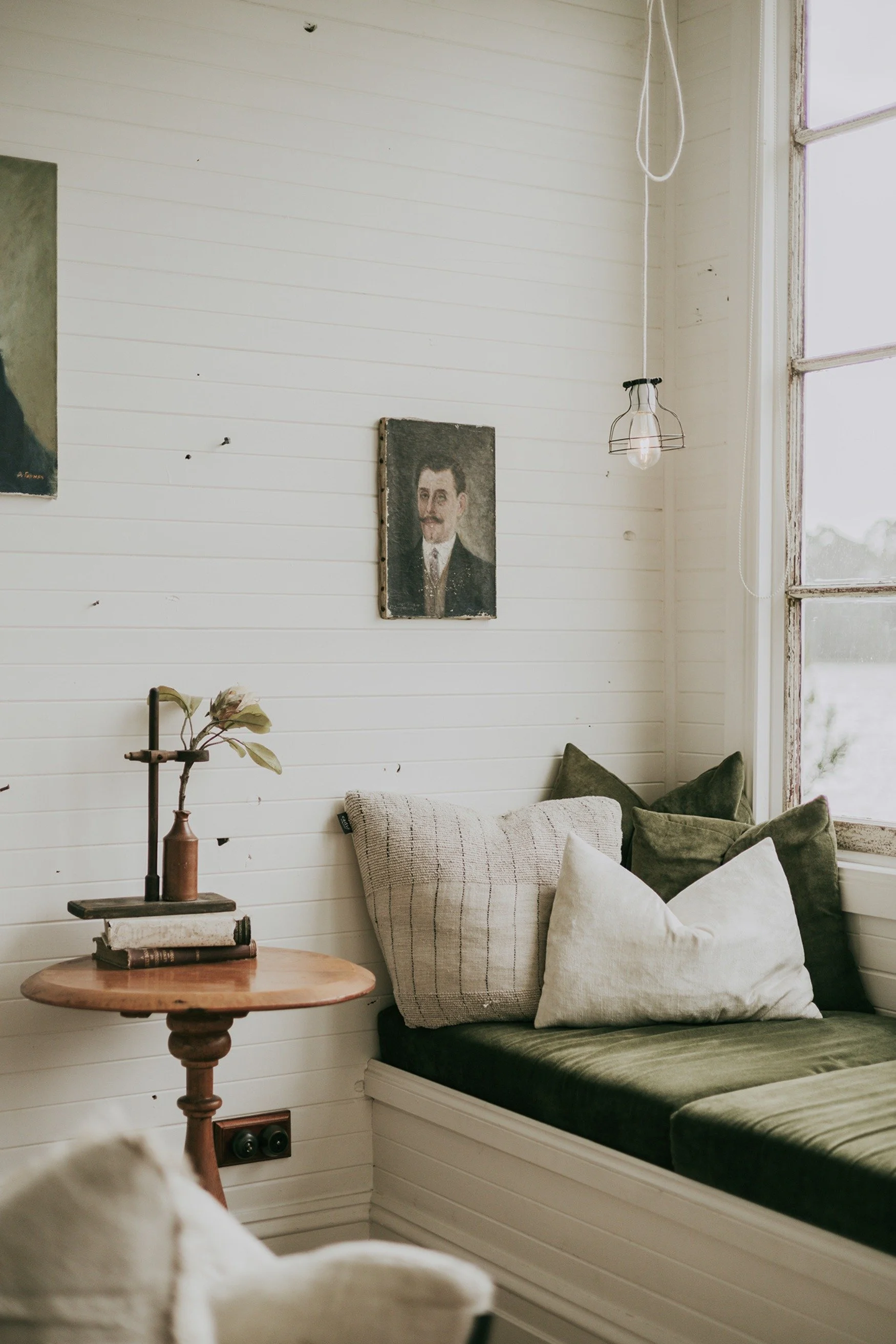 A living area with a day bed with cushions and a side table with books and a pot with a flower