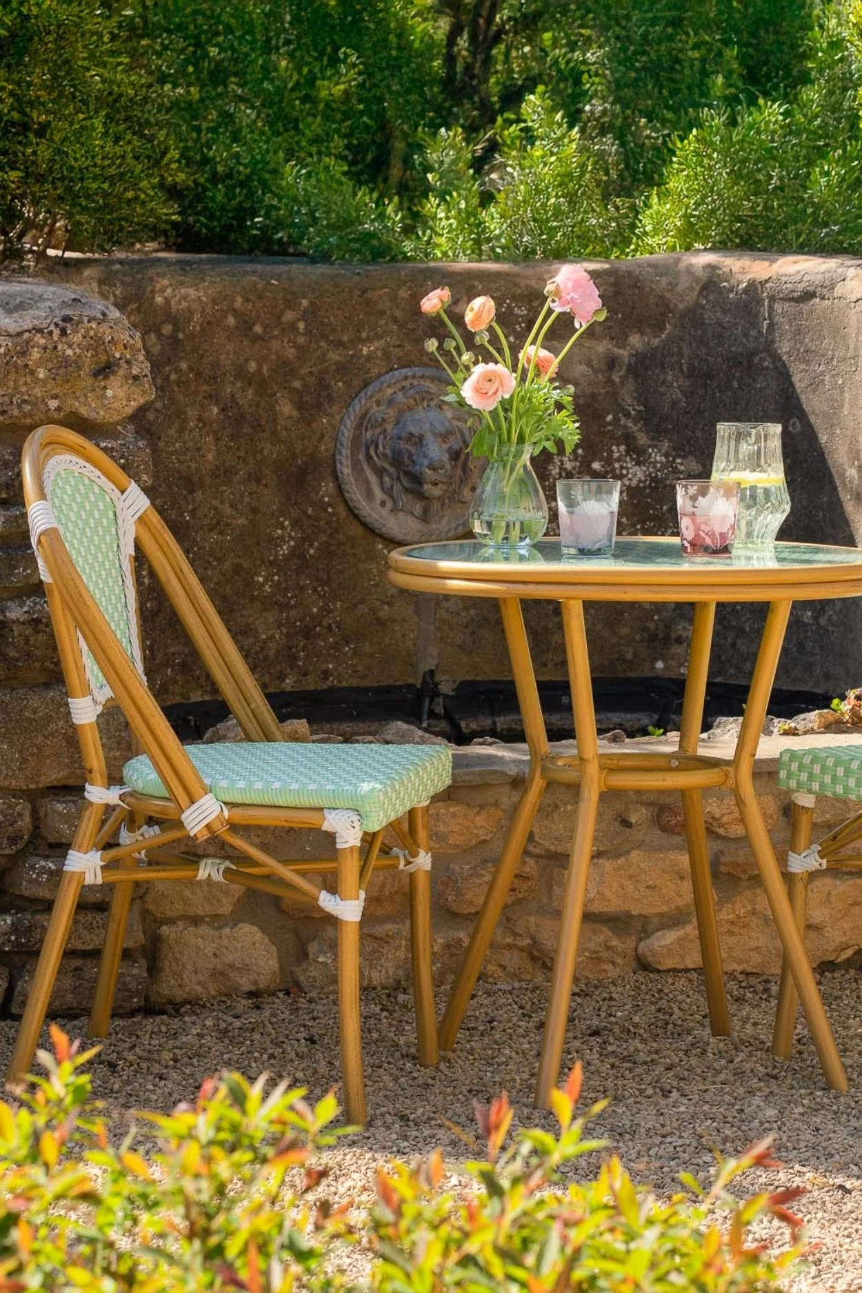 Mint green and white Parisian-style bistro table and chairs set in a gravel courtyard with flowers and drinks.